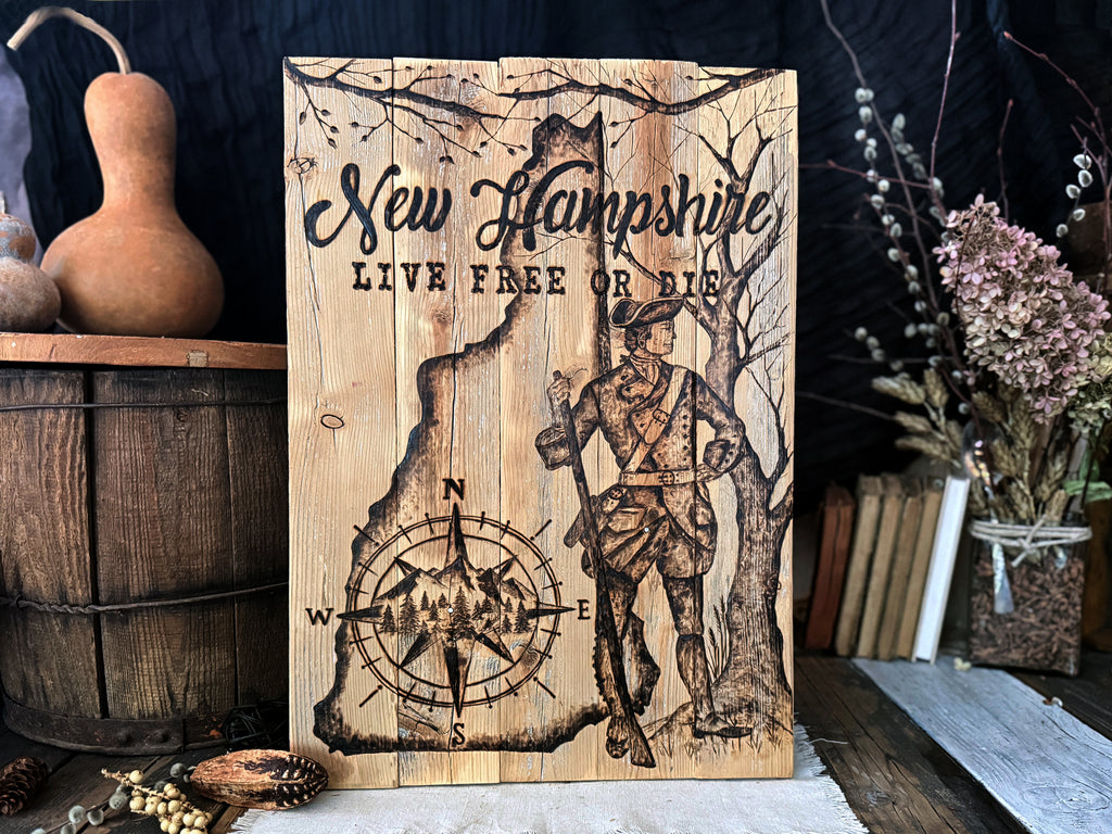 A rustic wooden sign featuring the text 'New Hampshire' and 'LIVE FREE OR DIE' with a hand drawn wood burned illustration of a colonial soldier, trees, and a compass rose. The sign is displayed on a rustic wooden surface surrounded by decorative props including a gourd, a wooden barrel, books, and dried flowers.