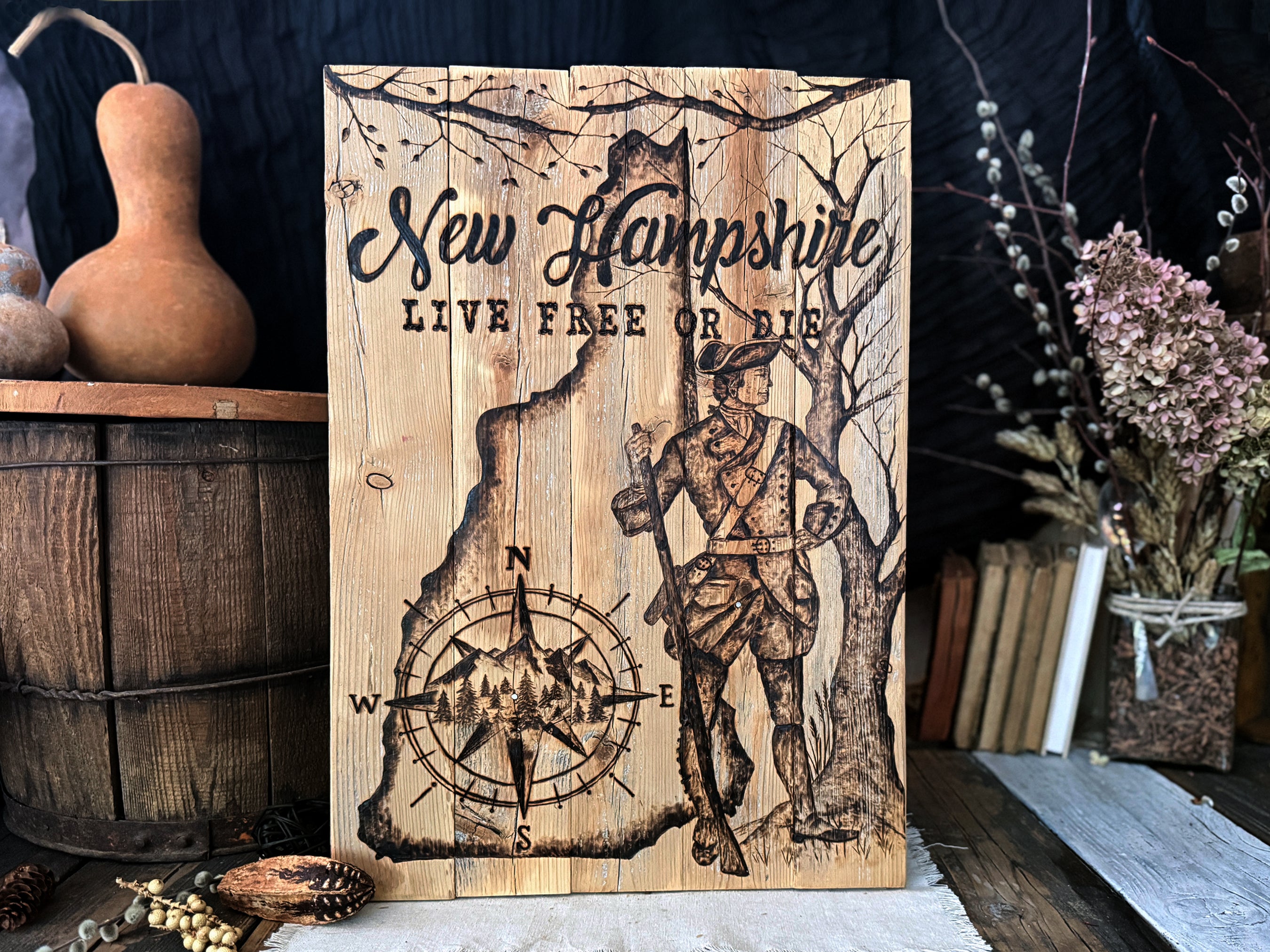 A rustic wooden sign featuring the text 'New Hampshire' and 'LIVE FREE OR DIE' with a hand drawn wood burned illustration of a colonial soldier, trees, and a compass rose. The sign is displayed on a rustic wooden surface surrounded by decorative props including a gourd, a wooden barrel, books, and dried flowers.