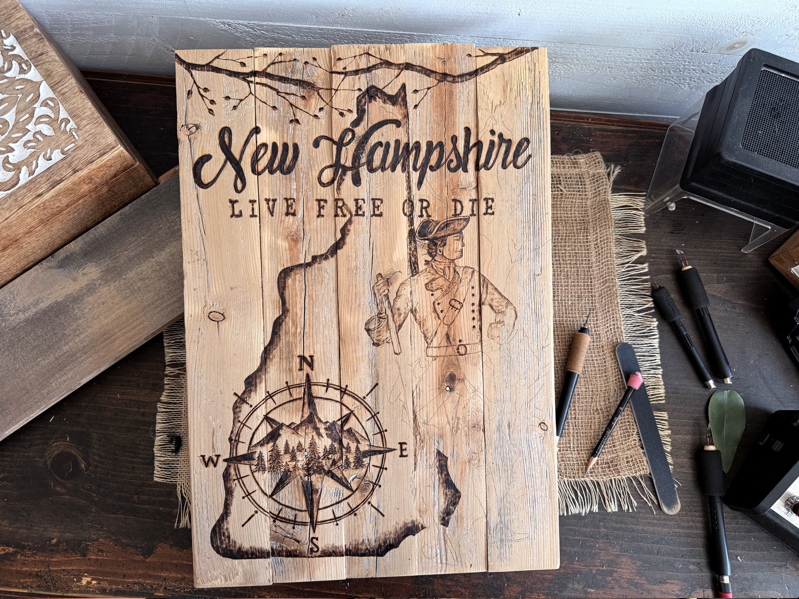 A rectangular wooden sign featuring a rustic engraved map of New Hampshire with the text 'New Hampshire' and 'LIVE FREE OR DIE,' plus a compass rose and figure illustration, displayed on a dark wooden surface with other wood and crafting items around it.