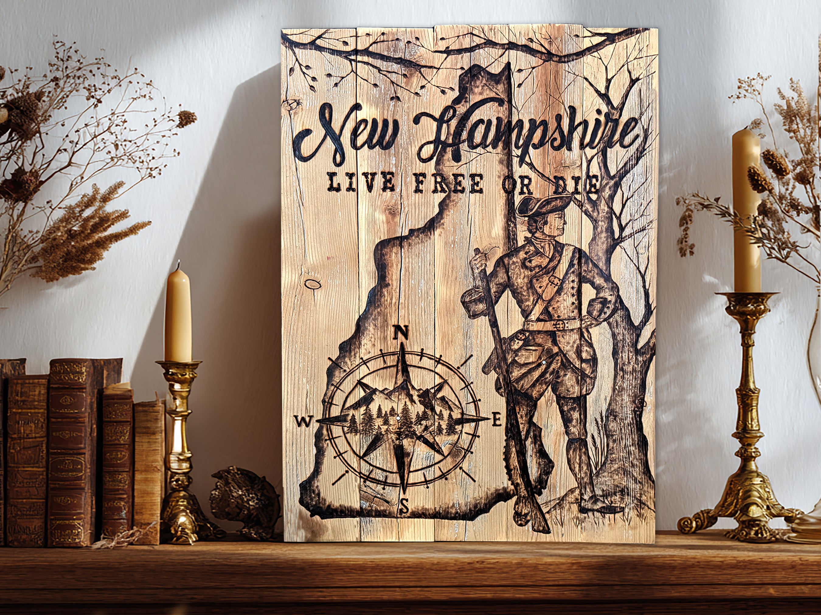 A rustic wooden sign featuring the text 'New Hampshire' and 'LIVE FREE OR DIE' with an wood burned hand drawn colonial soldier and compass rose design, displayed on a wooden shelf between books, dried flowers, and candlesticks.