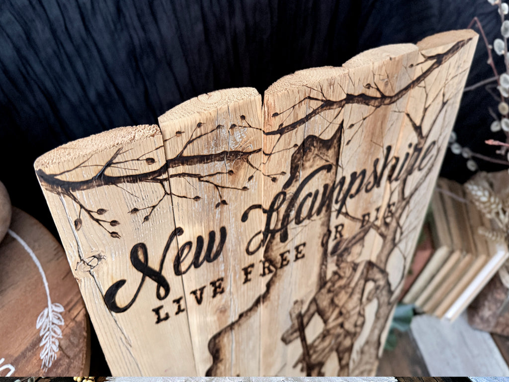 A close-up of the top boards of a rustic wooden sign featuring the words 'New Hampshire' and 'LIVE FREE OR FREEZE' in dark engraved lettering, with branch and tree illustrations across the light natural wood surface.