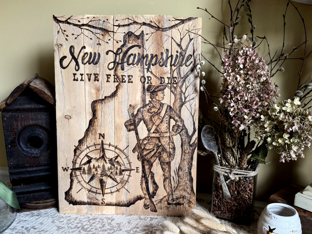 A rustic wooden sign featuring the text 'New Hampshire' and 'LIVE FREE OR DIE' with an wood burned hand drawn soldier and compass design, displayed on a tabletop with dried flowers and decorative objects in a styled interior setting.