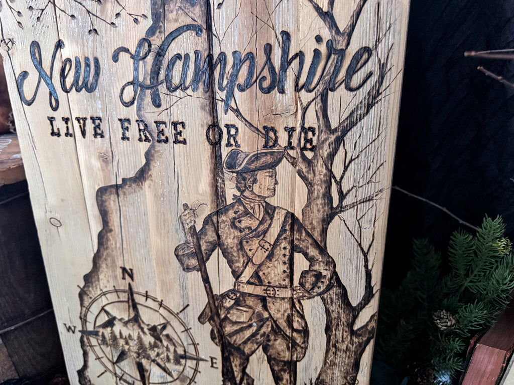 A rustic wooden sign featuring a hand drawn wood burned illustration of a Revolutionary War soldier, a compass rose, and the text 'New Hampshire' and 'LIVE FREE OR DIE' on a weathered wood background.