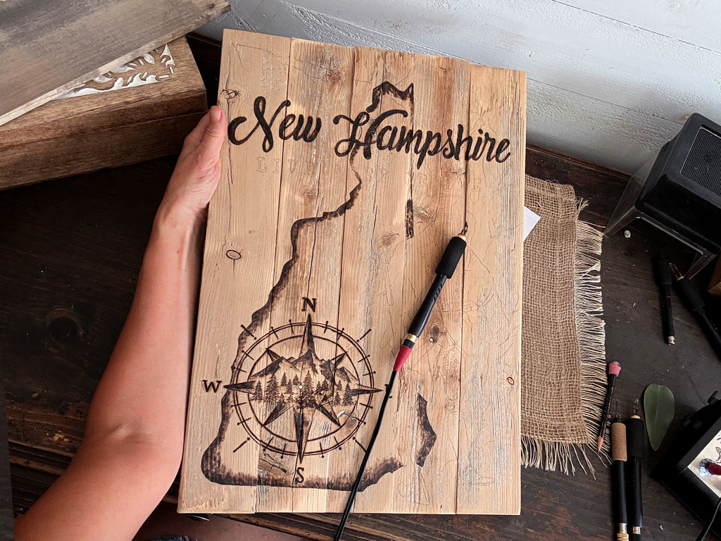 A hand holds a rustic wooden plaque featuring a hand drawn wood burned  map of New Hampshire with a compass rose design and the words 'New Hampshire' at the top. A pyrography pen rests on the wooden surface, with a dark wood tabletop and other craft items visible in the background.