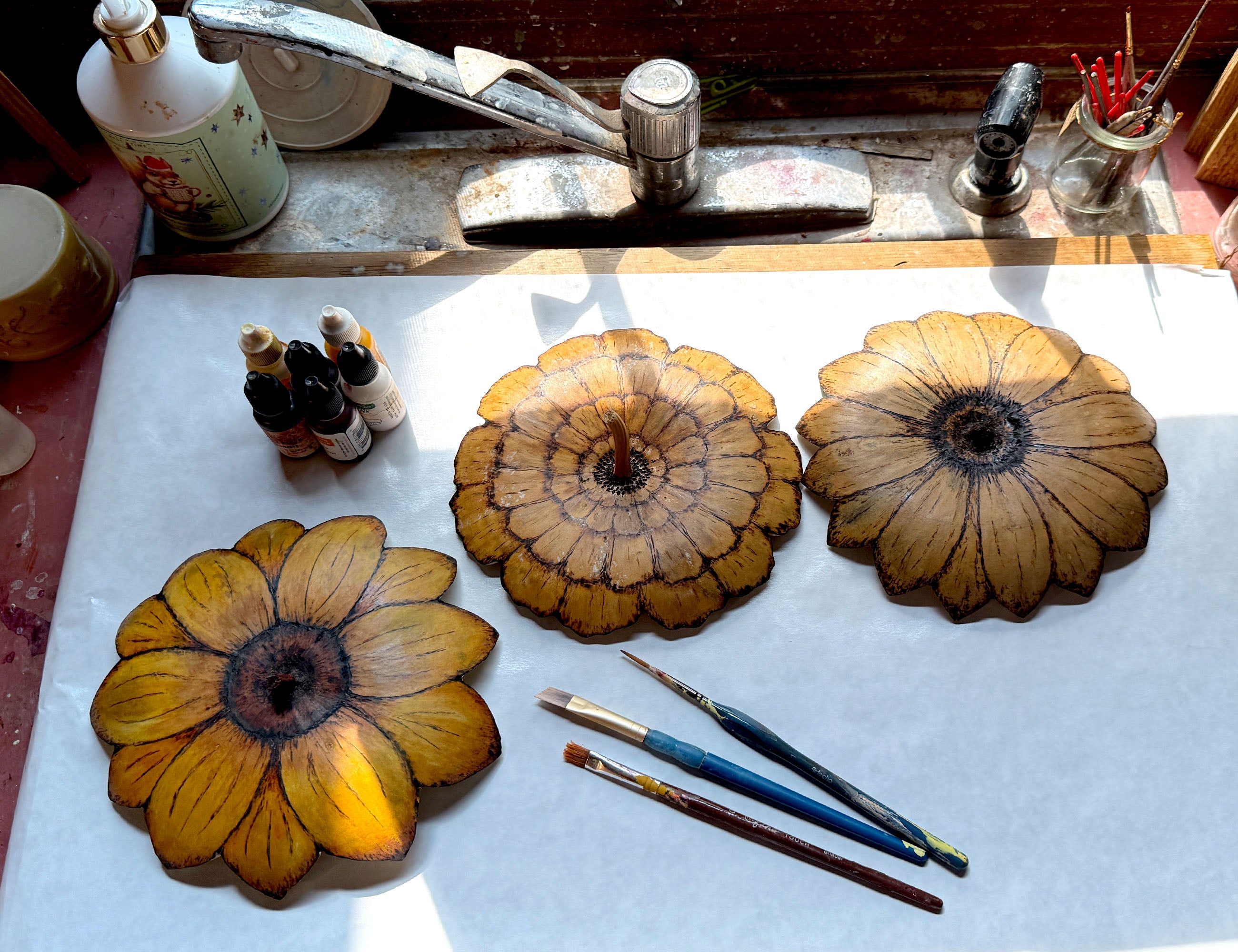 Three gourd lids with flower carvings on a white surface with painting tools and a sink in the background.