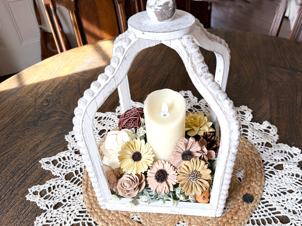 Decorative white lantern with a candle and flowers on a wooden table.