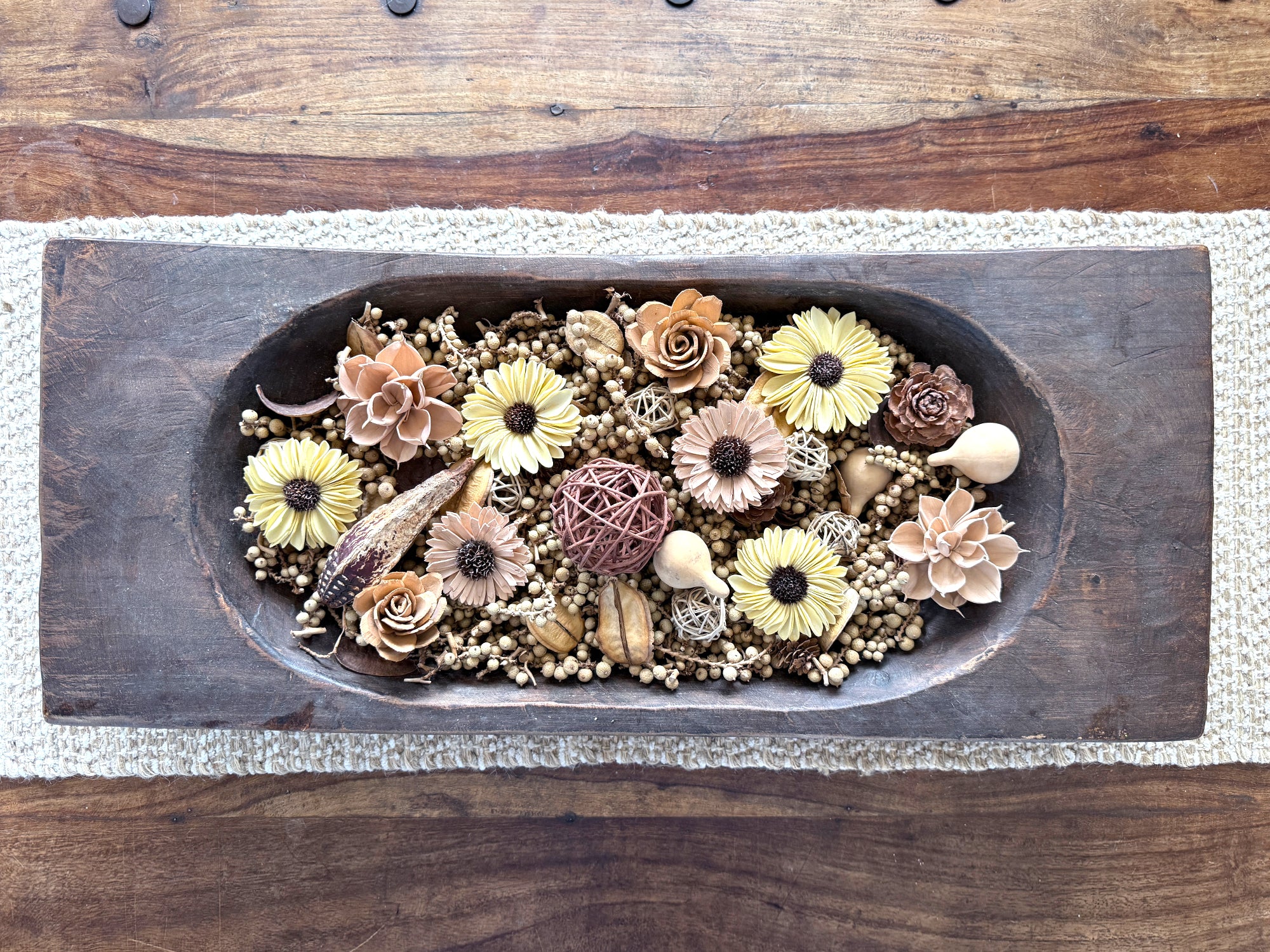 Decorative dough bowl arrangement of flowers and natural elements in a large wooden dough bowl on a coffee table.