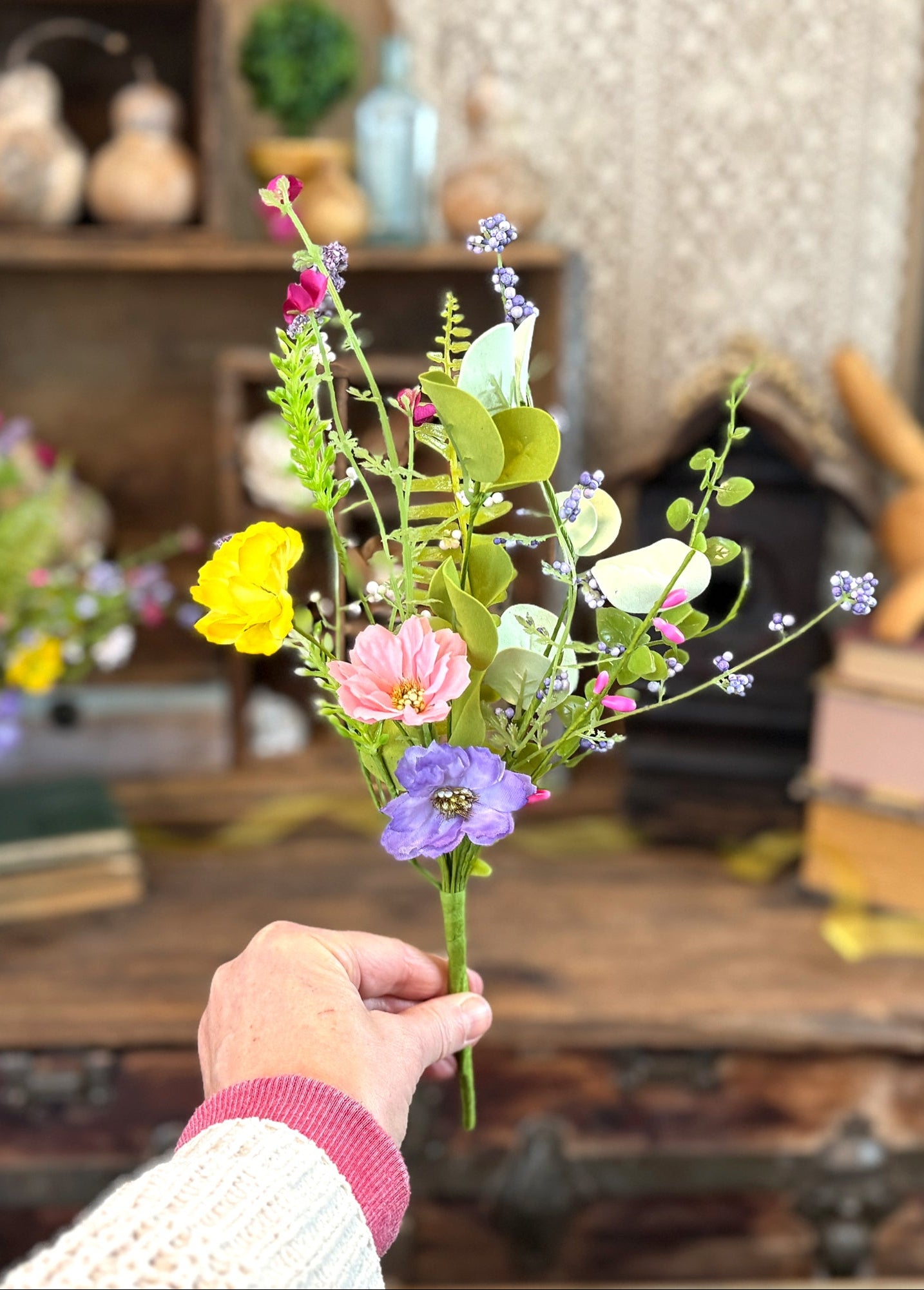 Person holding a small bouquet of artificial flowers in a room with books and decorative items.