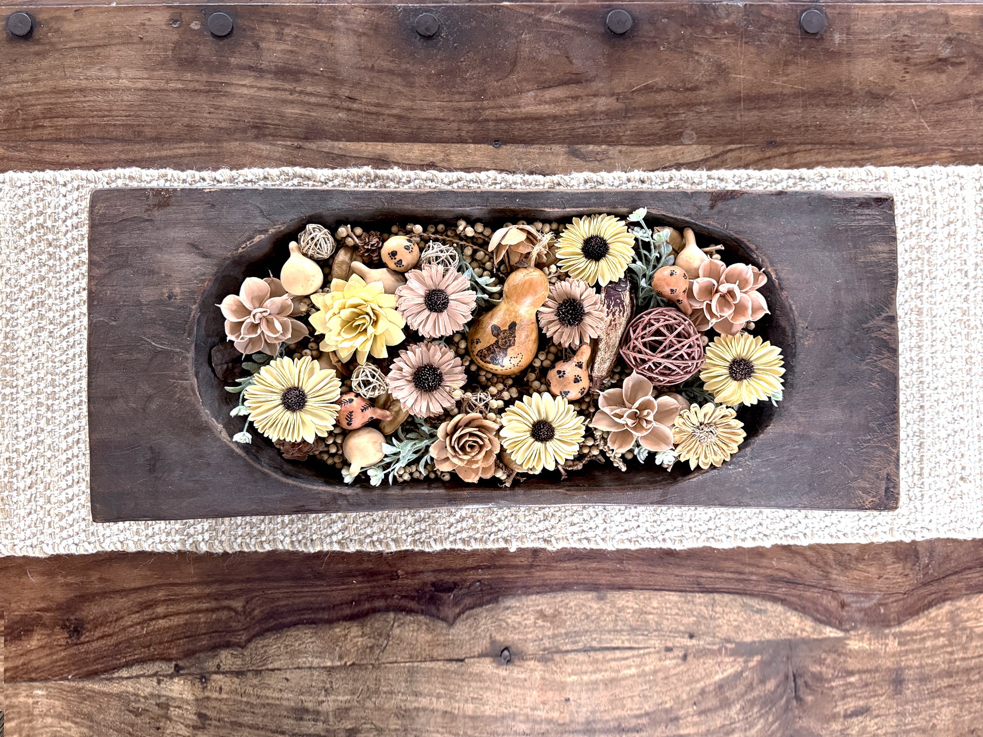 Decorative arrangement of flowers and gourds in a wooden dough bowl on a wooden coffee table.