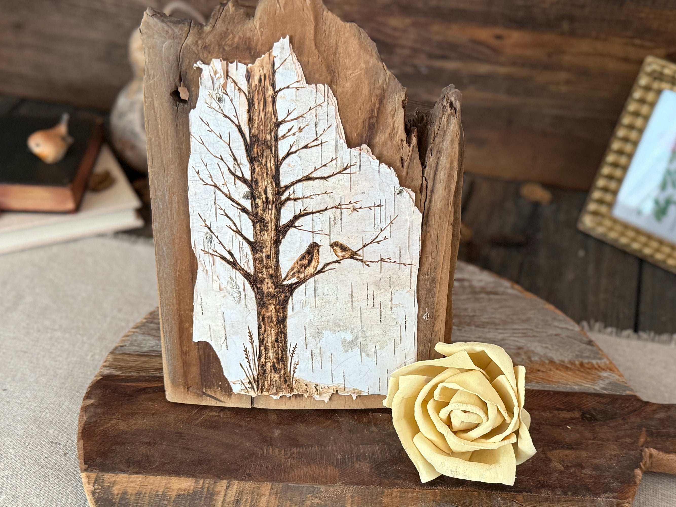 Wooden plaque with hand drawn tree and bird design next to a yellow rose on a rustic background