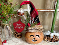 Decorative gnome with a red hat and brown body, surrounded by festive elements like pine cones and a wreath.