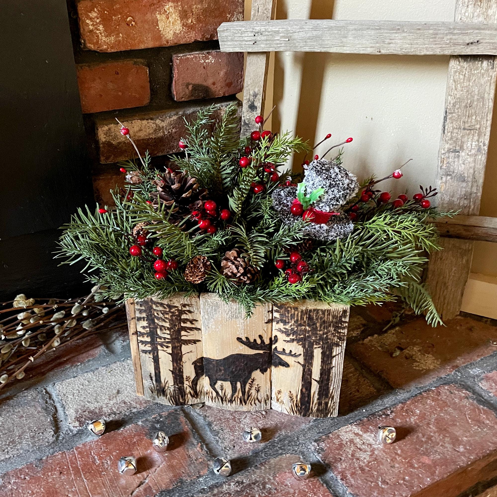 Decorative Christmas arrangement with greenery, red berries, and a wooden box with deer design on a brick surface.