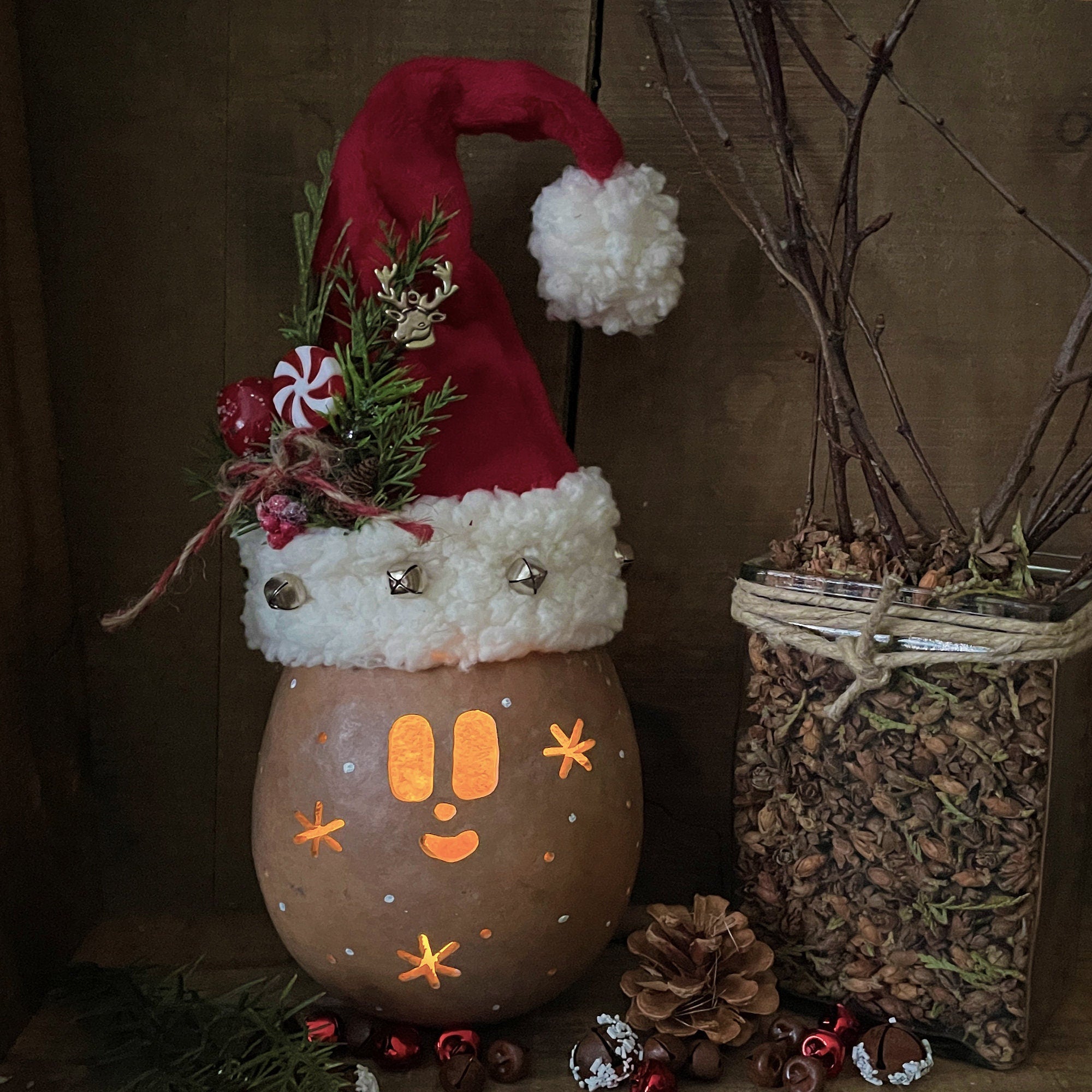 Decorative gourd with a Santa hat and face, surrounded by pinecones and a jar of dried plants on a brown background.