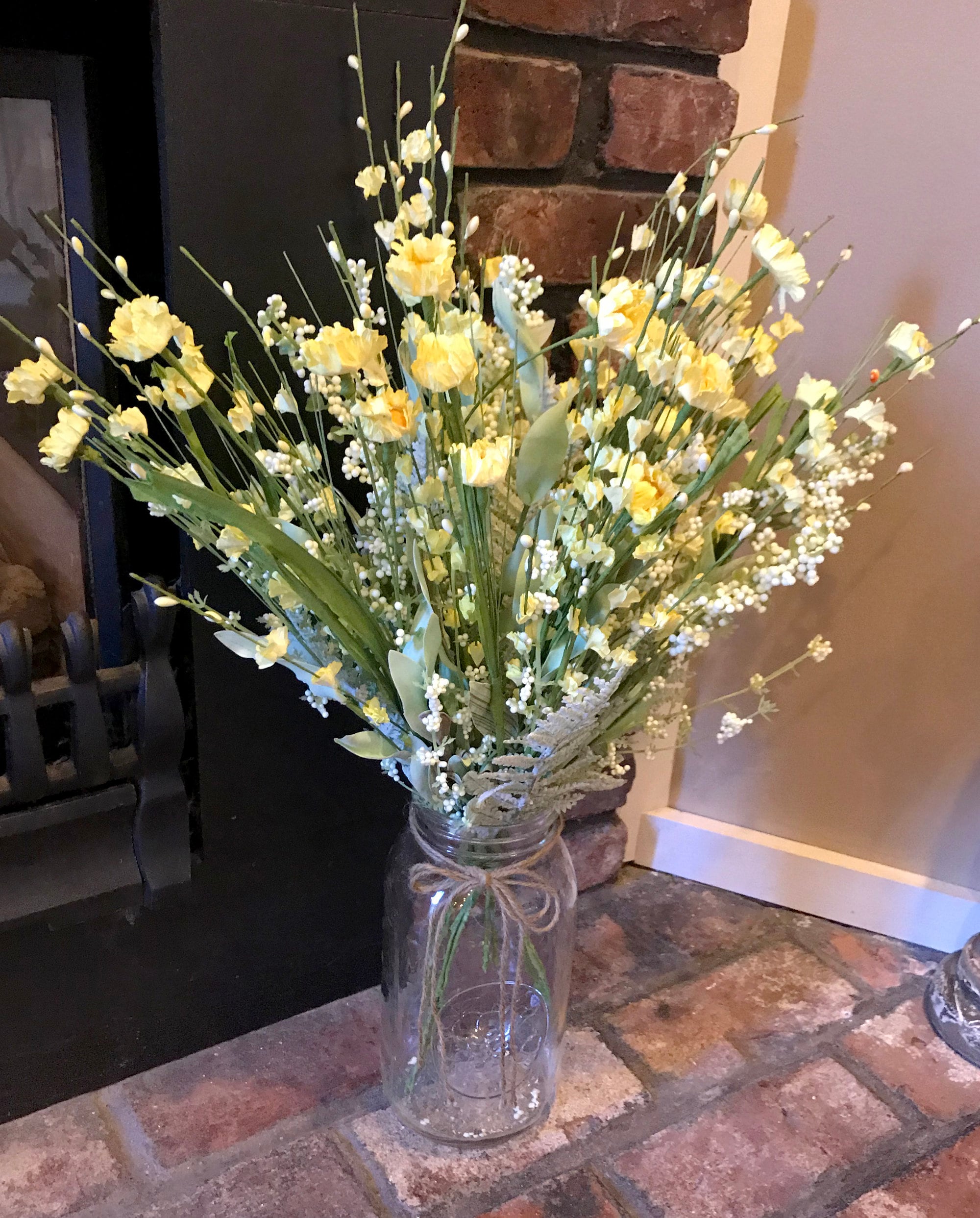 Bouquet of yellow flowers in a glass jar on a stone surface with a fireplace in the background.