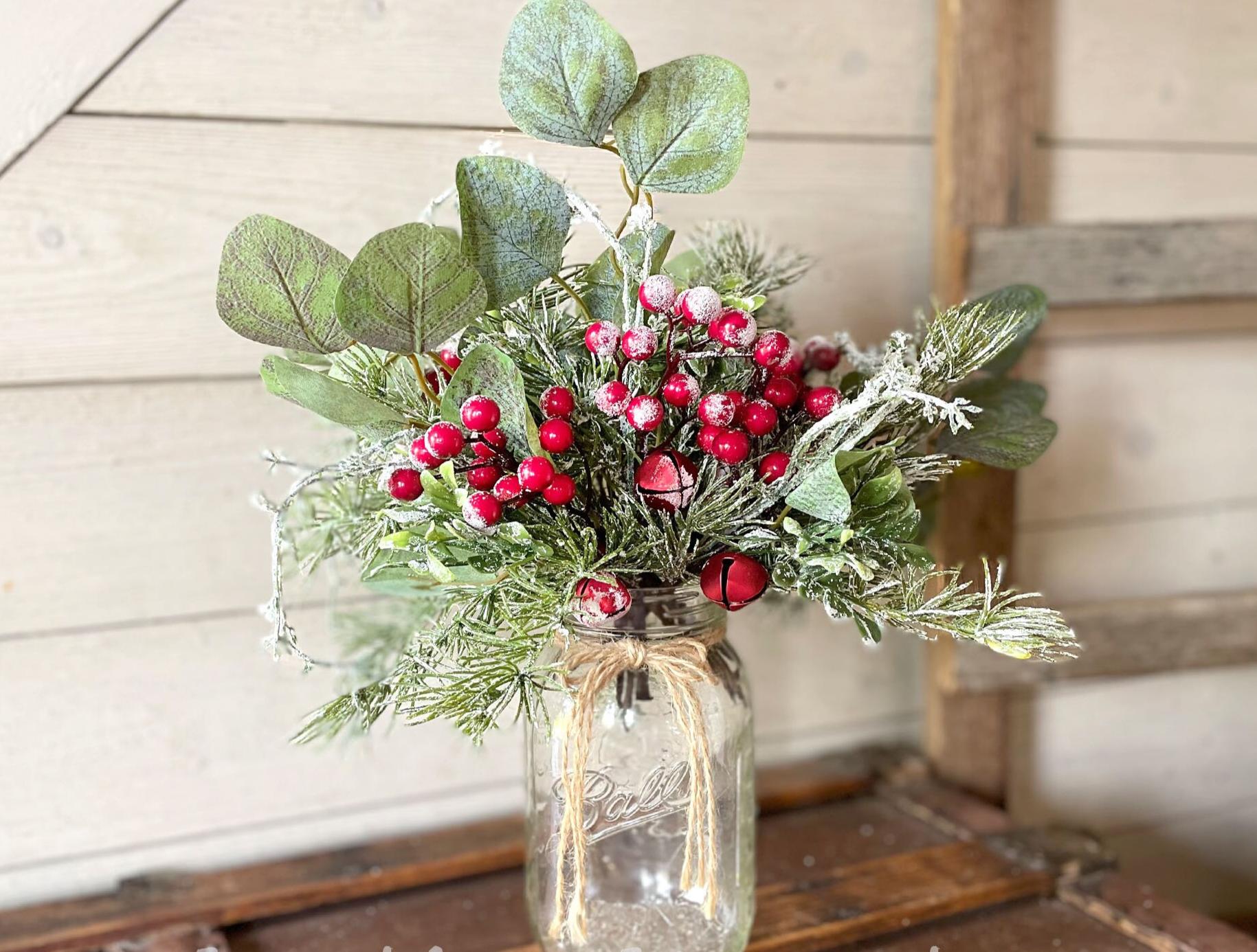 Bouquet of red berries and green leaves in a glass jar on a wooden surface.