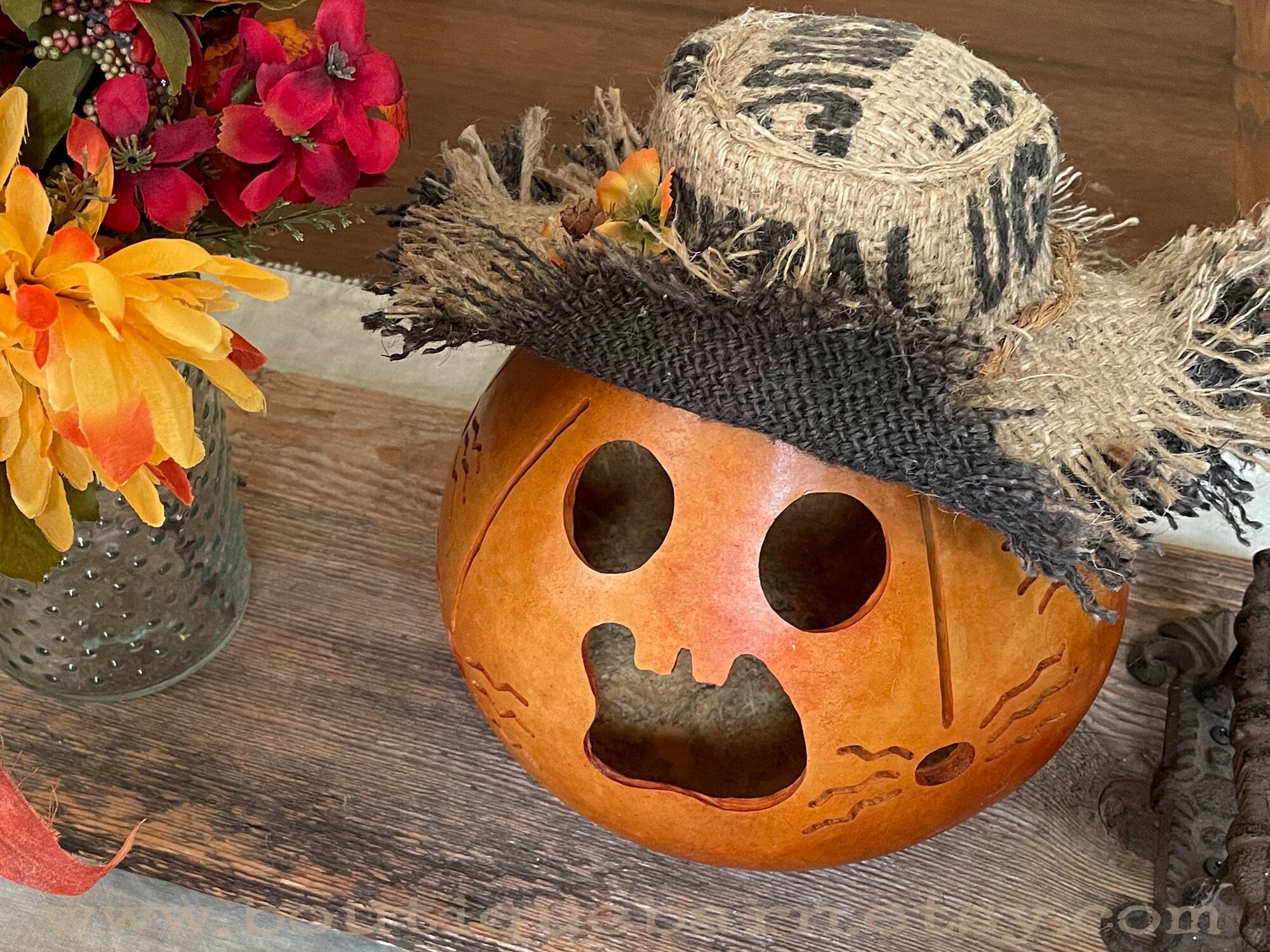 Decorative pumpkin with a straw hat on a wooden surface with flowers in the background.