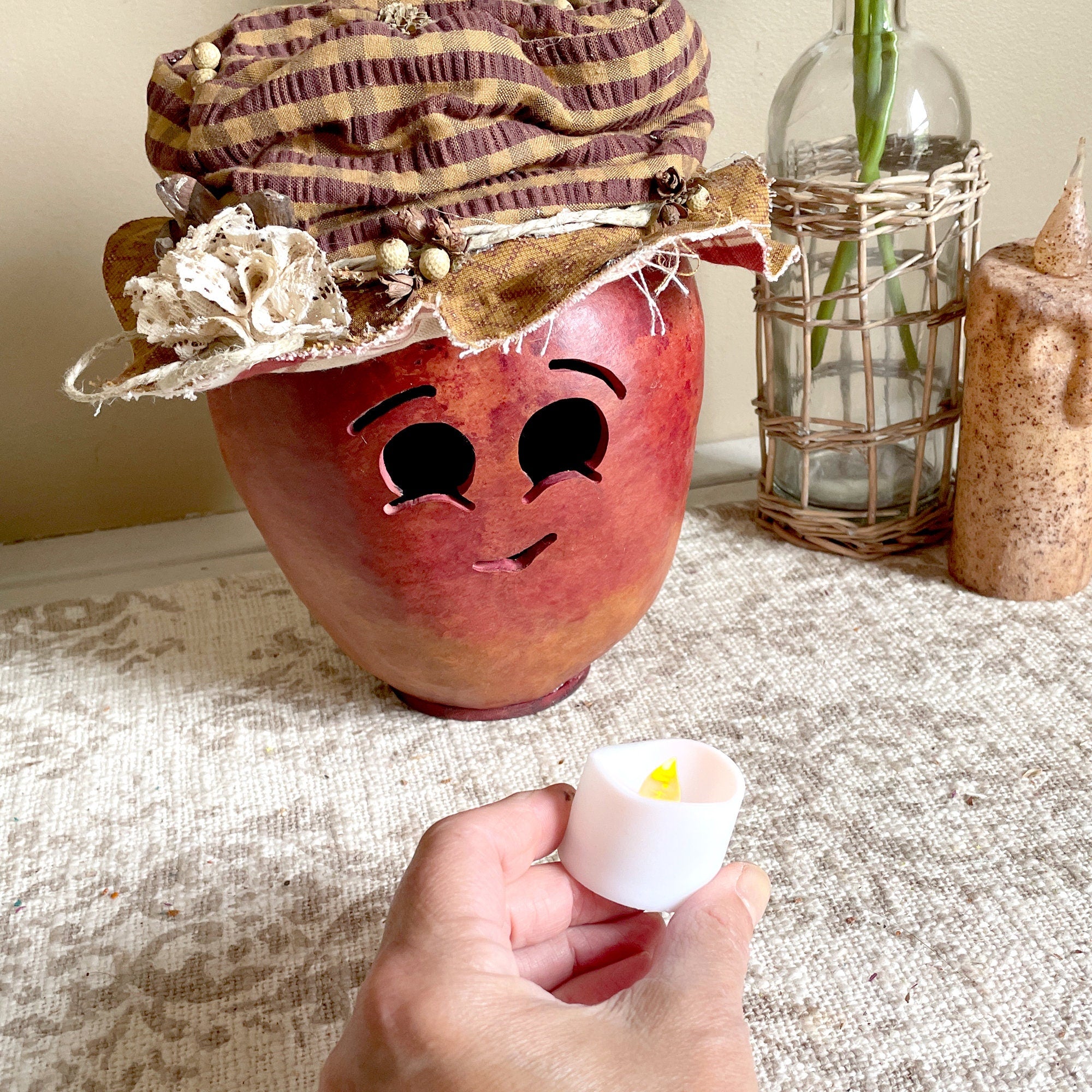 Decorative apple-shaped lantern with a face, held by a hand with a small white candle inside, on a textured surface.