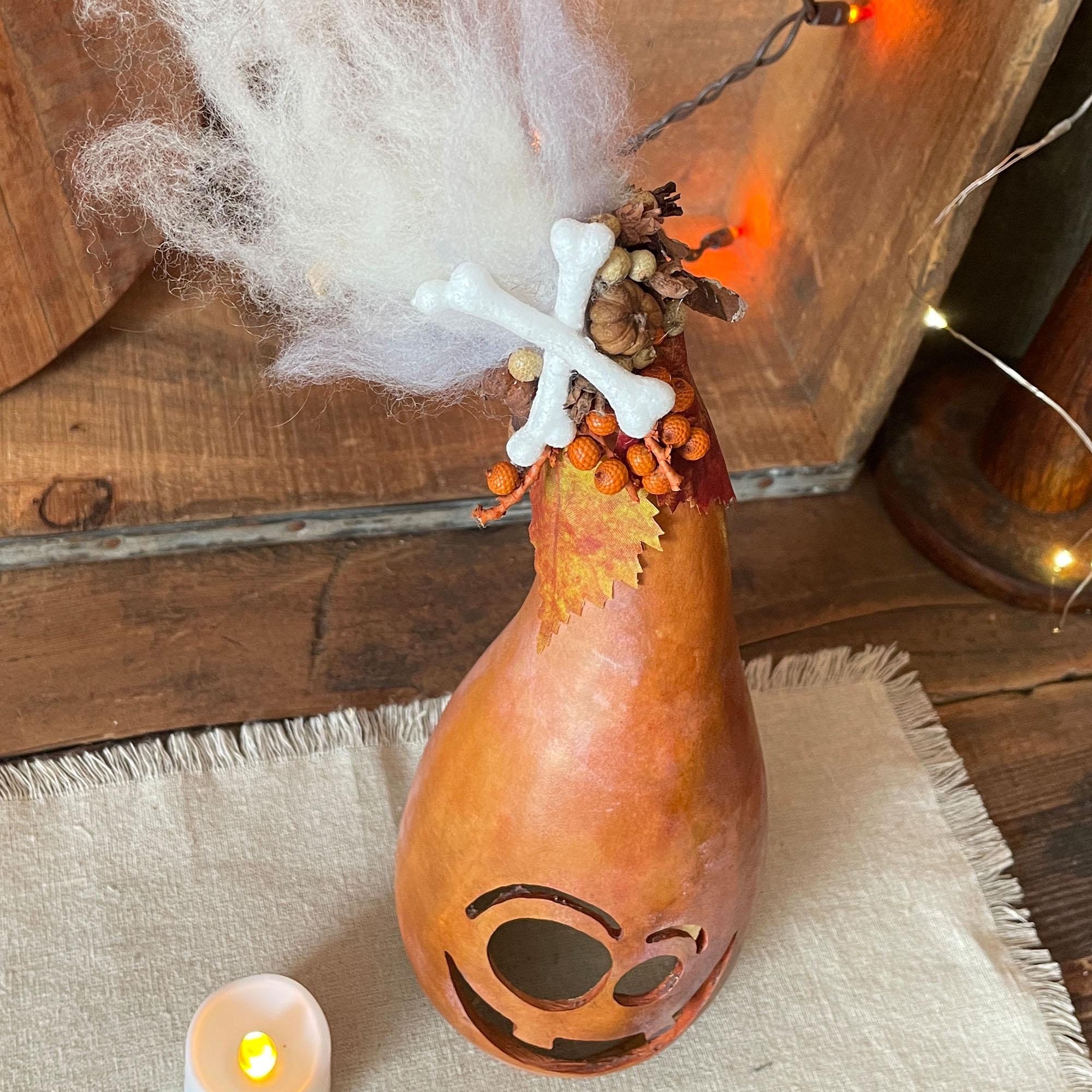 Decorative gourd with face and autumn leaves on a wooden surface with a candle and string lights.