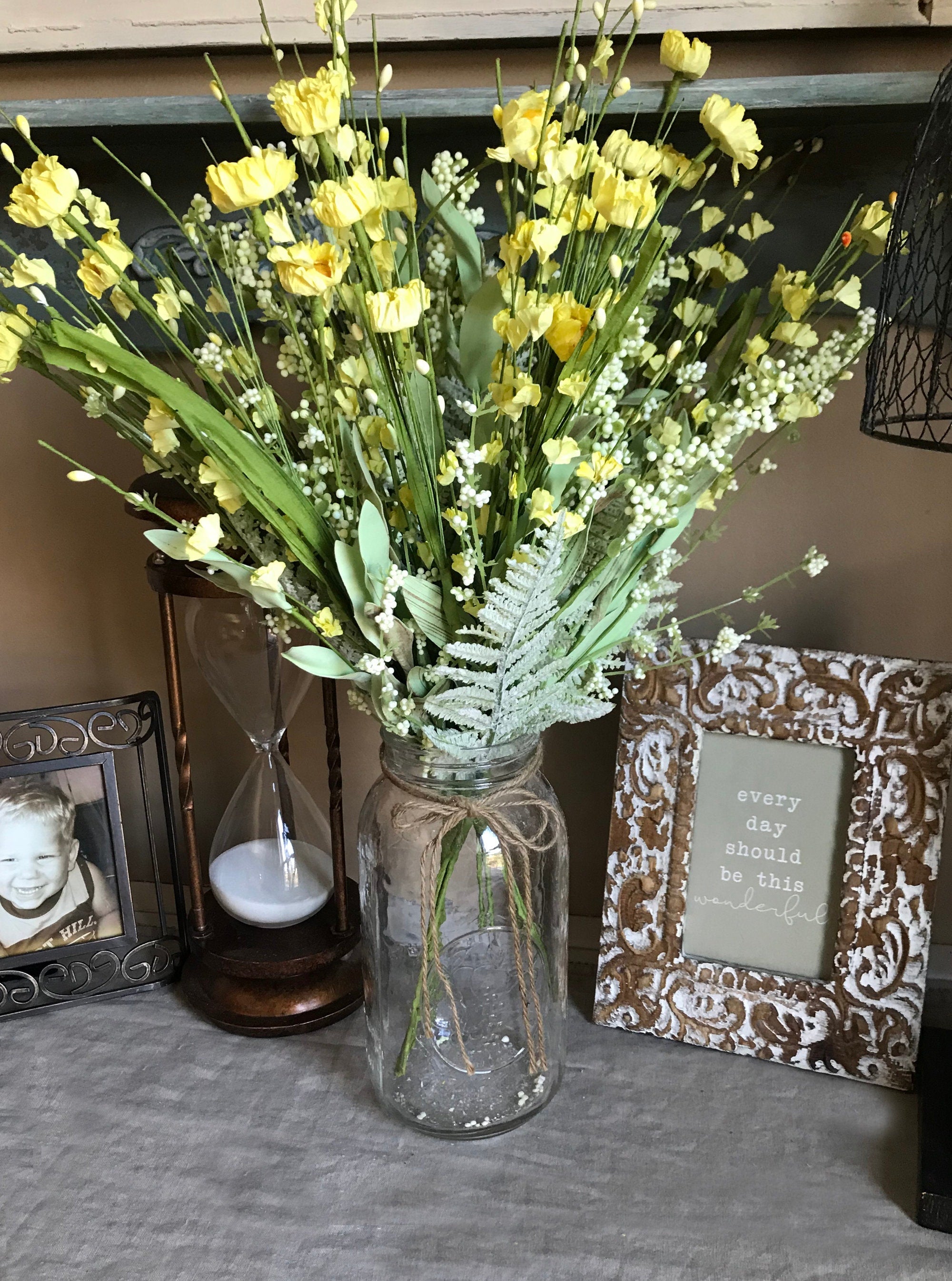 Bouquet of yellow flowers in a glass jar on a surface with decorative items in the background.