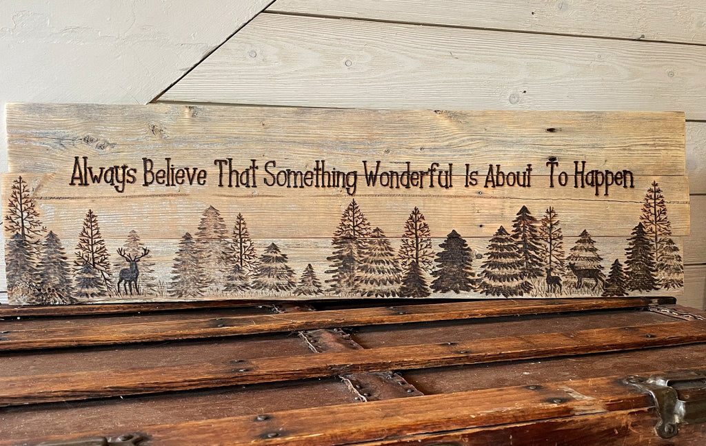 Wooden sign with engraved trees and text 'Always Believe That Something Wonderful Is About To Happen' on a wooden surface.