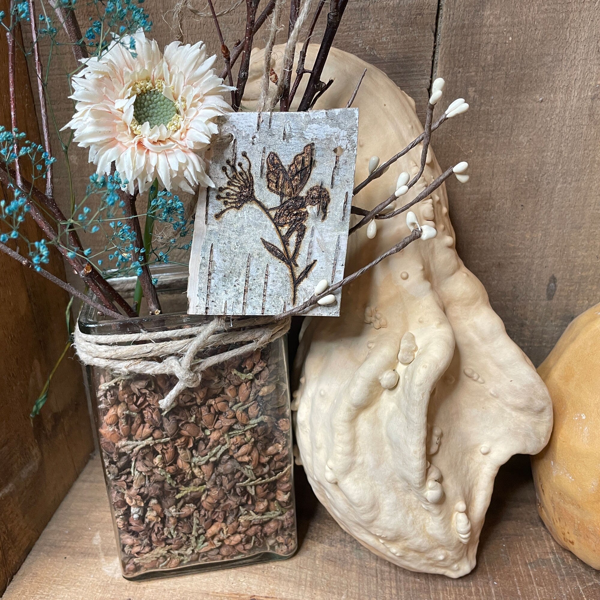 Decorative arrangement with flowers, branches, and a small wooden bark tag on a textured surface.