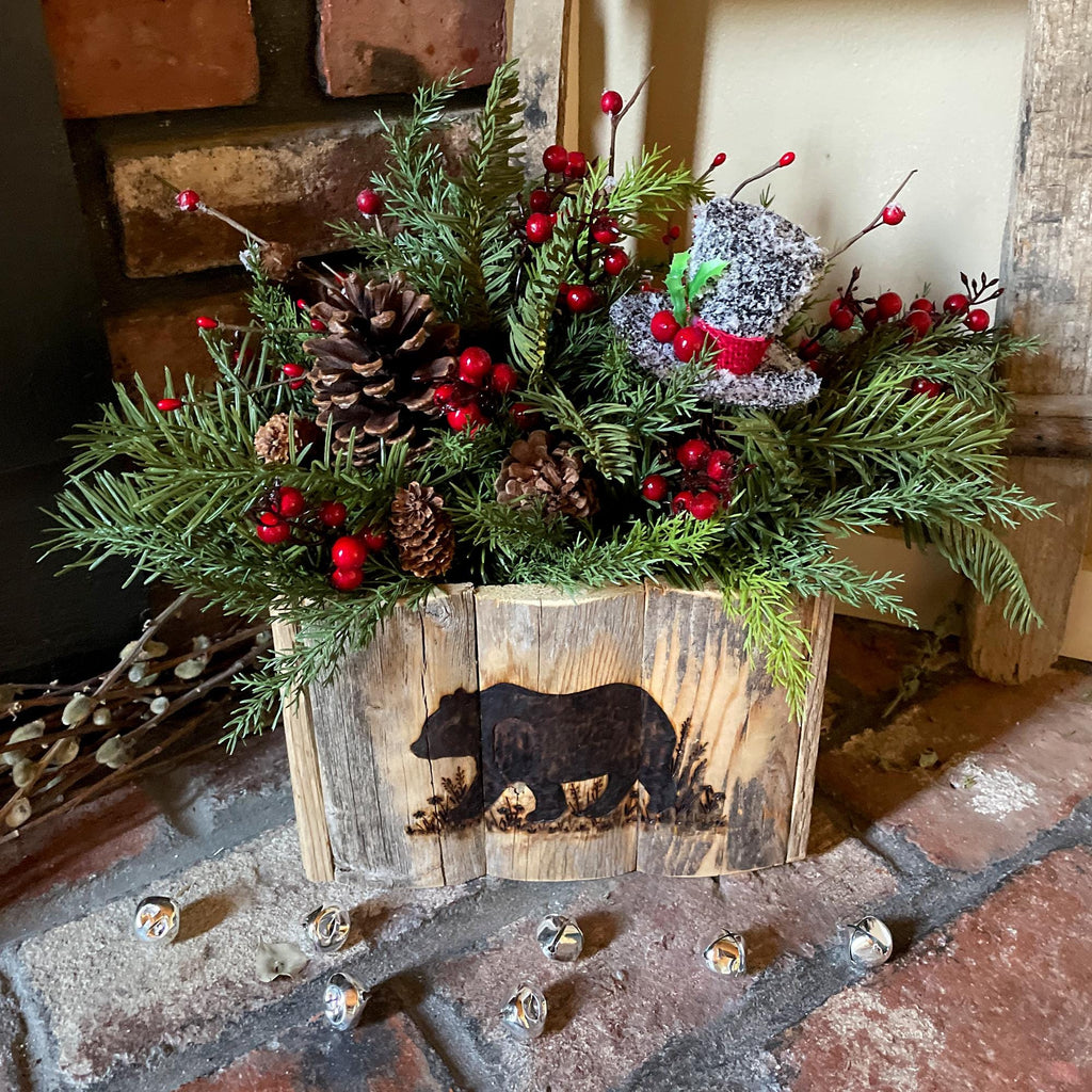 Decorative holiday arrangement with greenery, pinecones, and red berries in a wooden box with bear design on a stone surface.