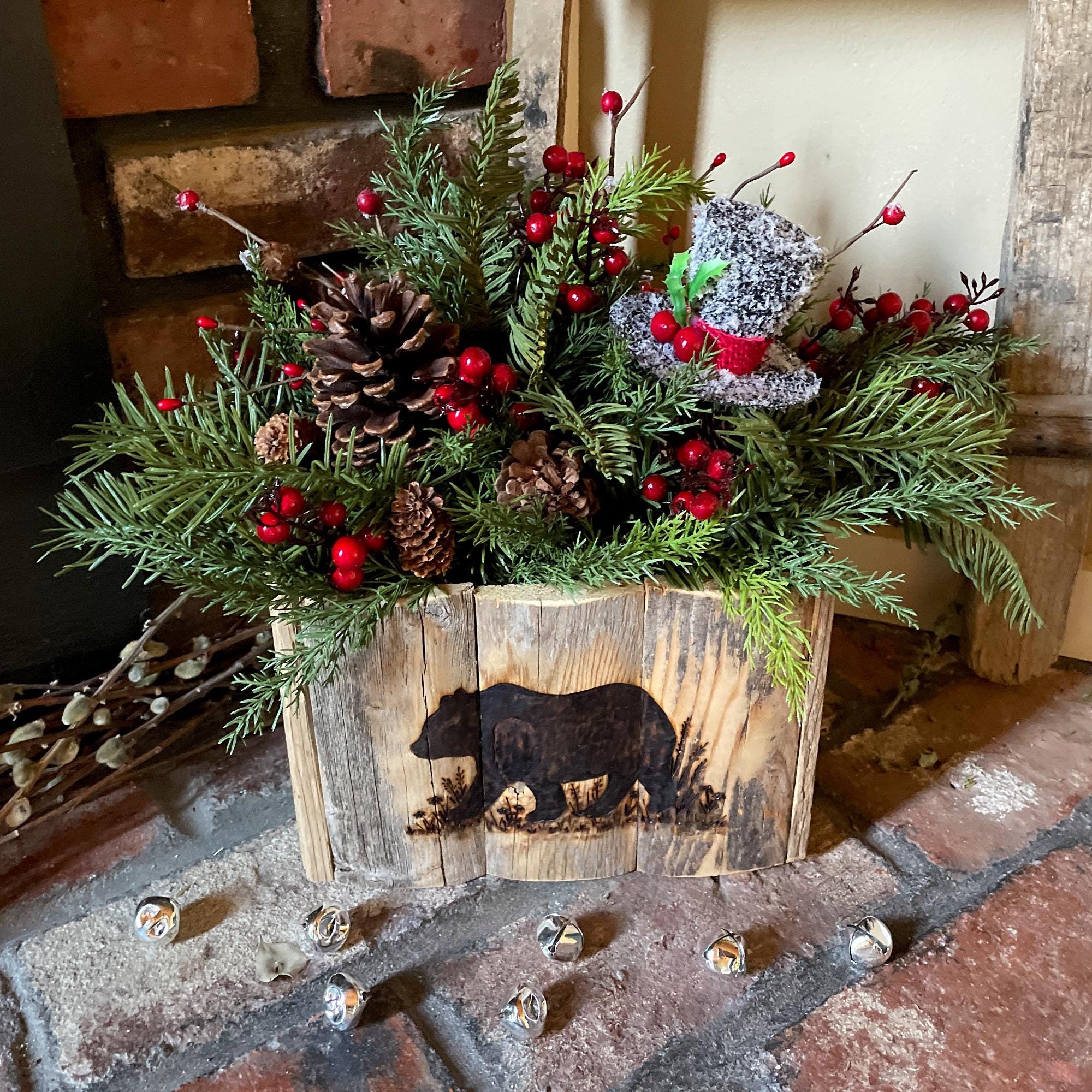 Decorative holiday arrangement with greenery, pinecones, and red berries in a wooden box with bear design on a stone surface.