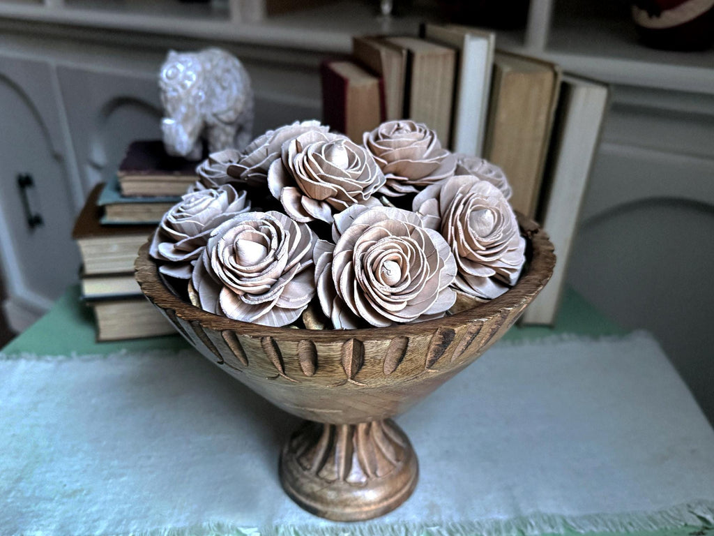 Decorative wooden rose arrangement in a pedestal bowl on a table with books and a decorative object in the background.