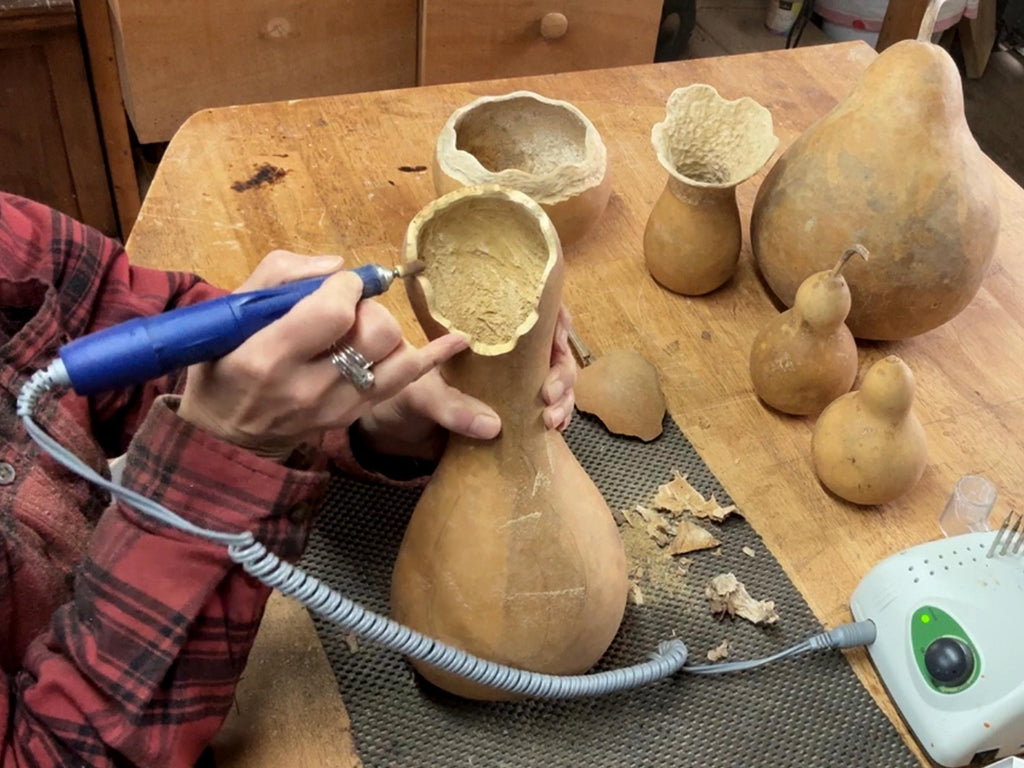 Person using a tool to work on a gourd on a wooden table with other gourds.
