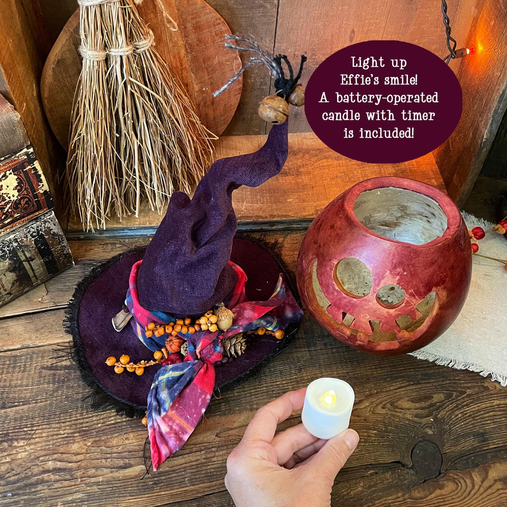 Decorative witch hat with a pumpkin lantern and battery-operated candle on a wooden surface.