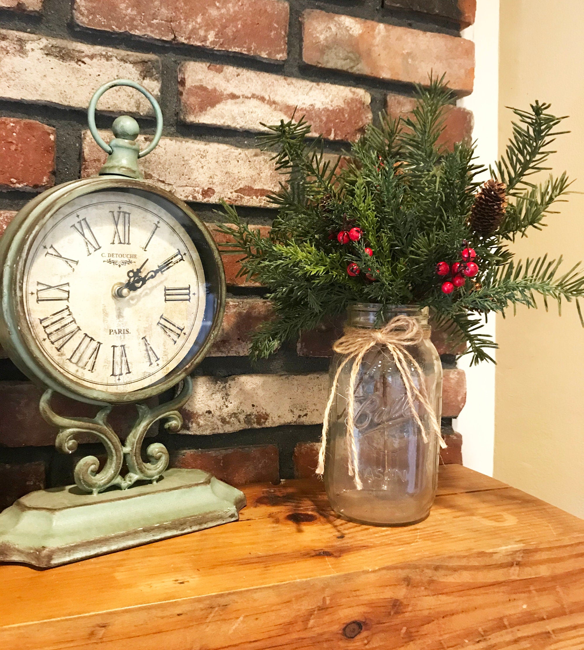 Decorative clock and jar with greenery on a wooden surface against a brick wall.