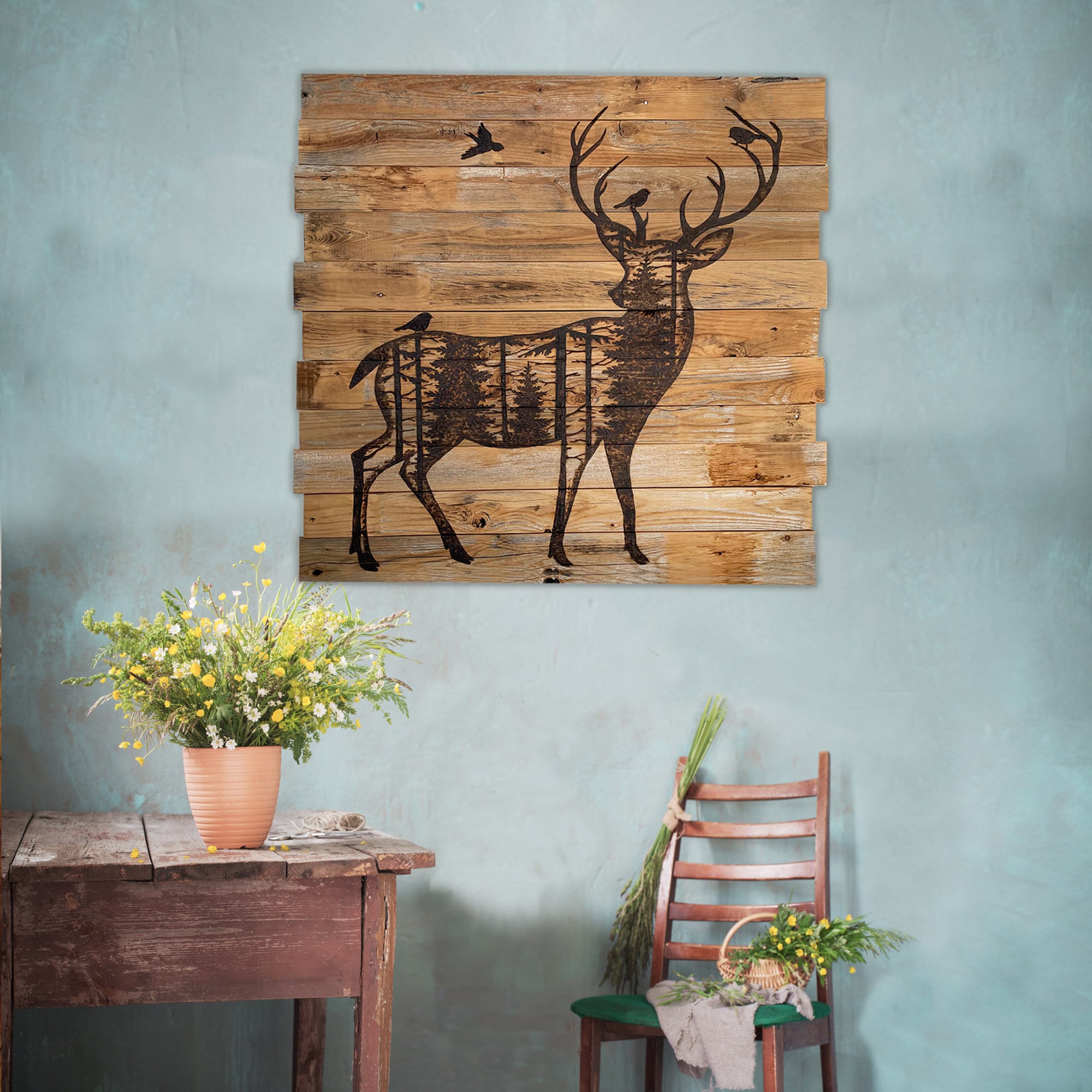 Wooden wall art with deer silhouette on a light blue wall, table with flowers, and chair with plants.