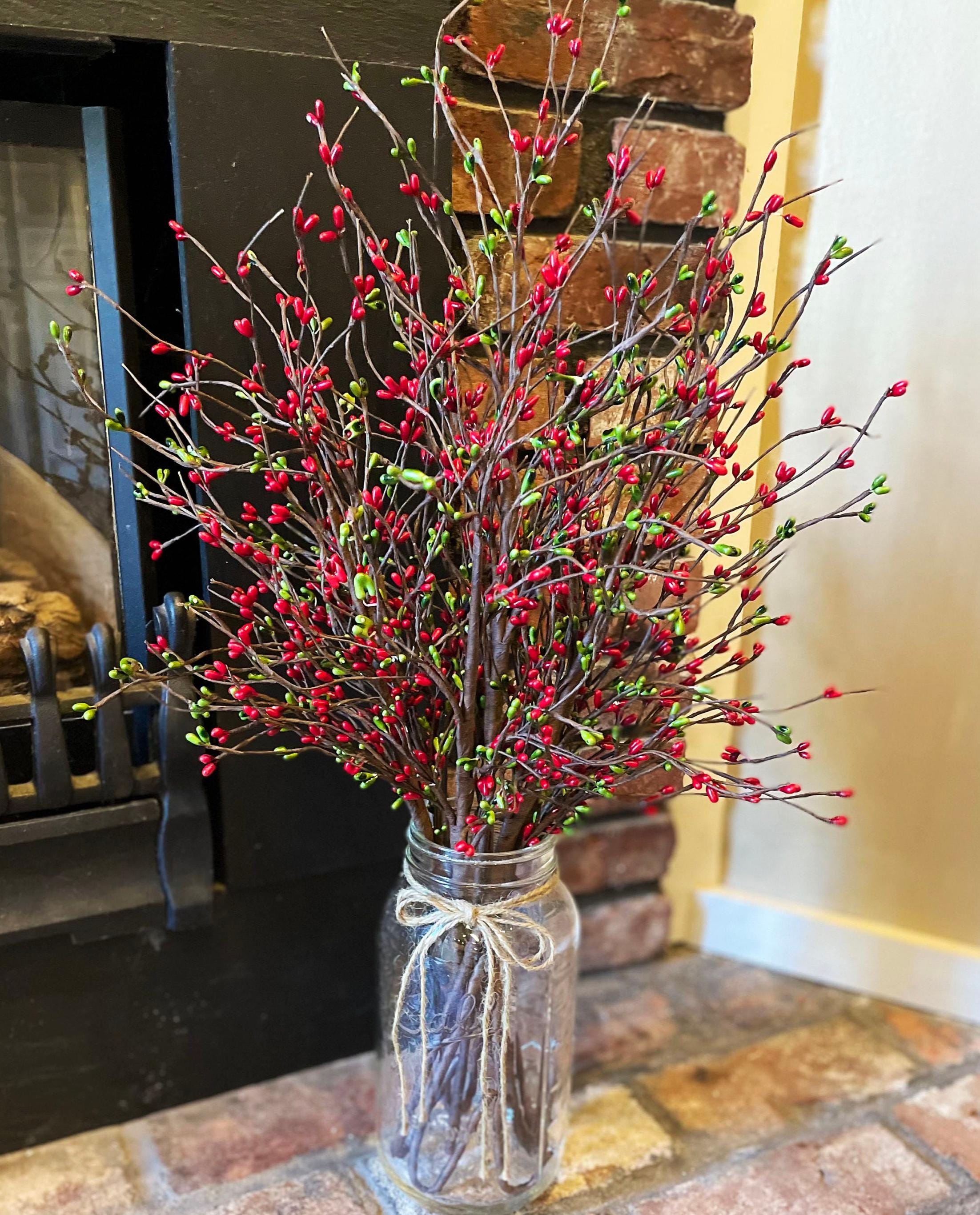 Decorative arrangement of red berries on branches in a clear glass jar with a white ribbon, placed on a stone surface.