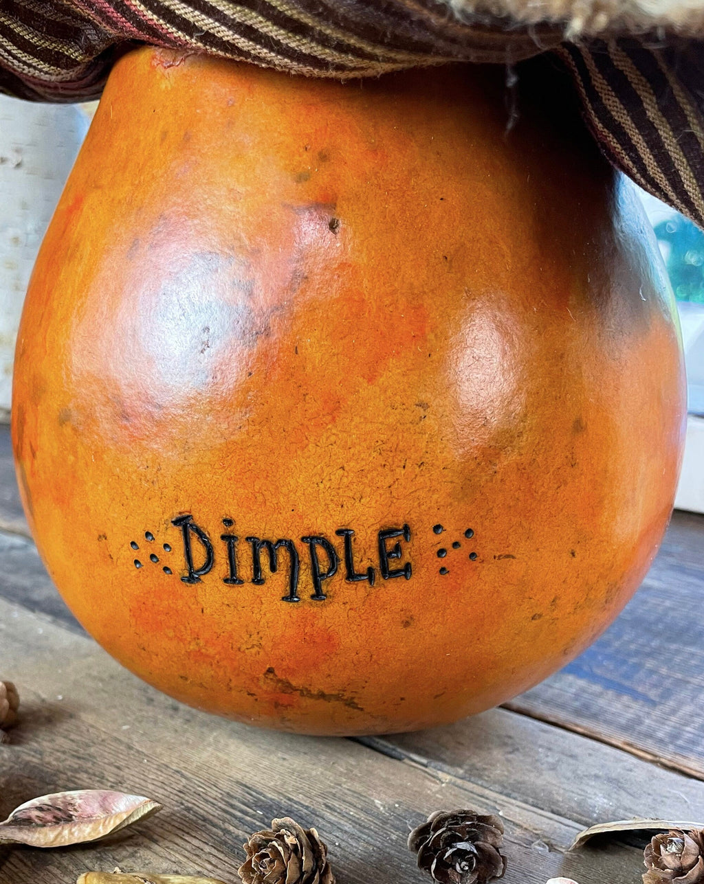 Decorative pumpkin with 'Dimple' branding on a wooden surface with pine cones.