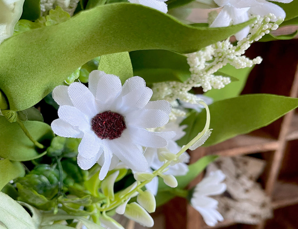Close-up of a white artificial flower with a brown center surrounded by green leaves and other flowers.
