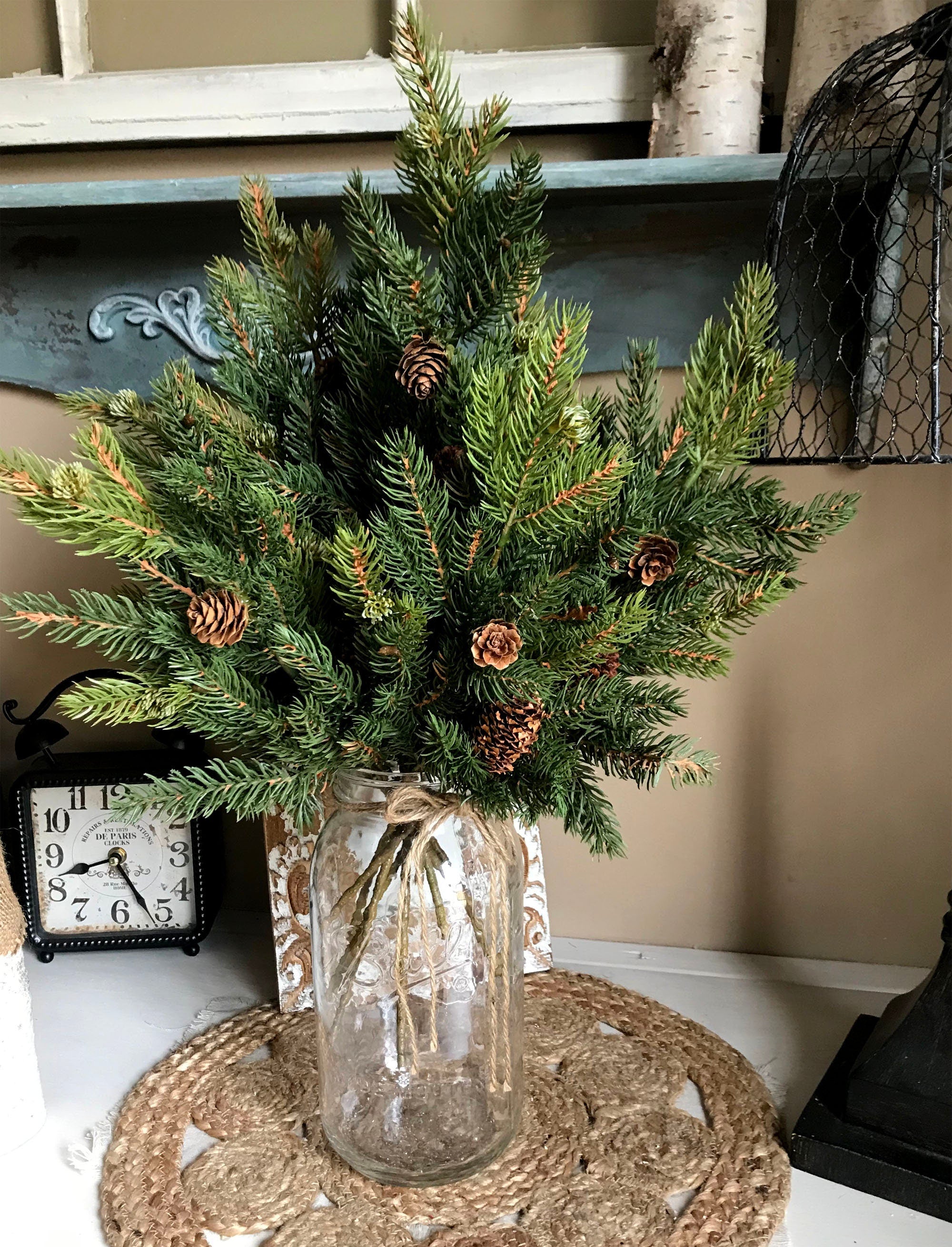 Vase with greenery and pinecones on a table with a clock in the background
