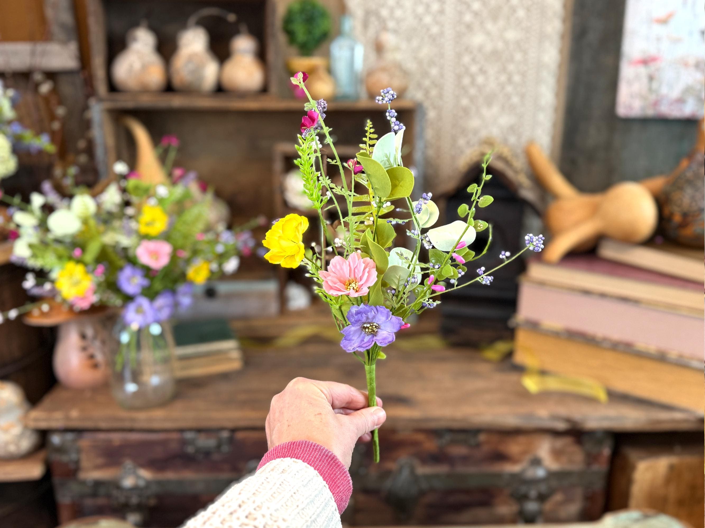 Person holding a small bouquet of artificial flowers in a room with books and decorative items.