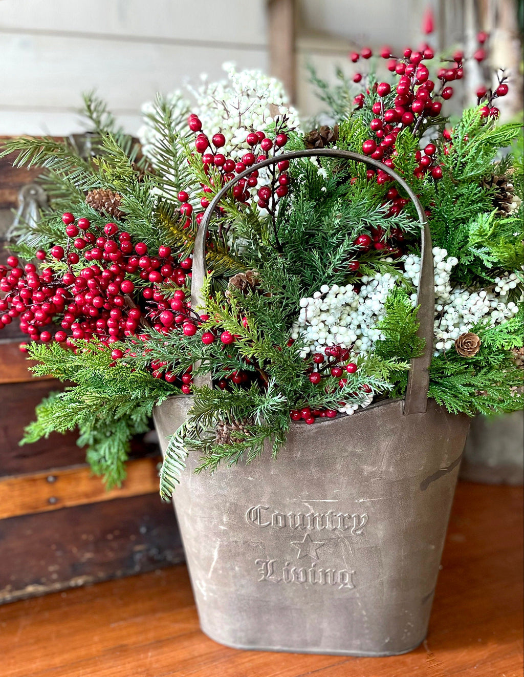 Decorative arrangement of greenery with red berries in a metal bucket on a wooden surface.