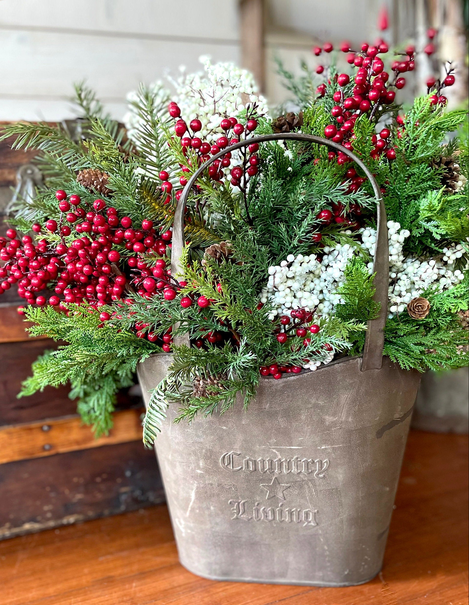 Decorative arrangement of greenery with red berries in a metal bucket on a wooden surface.