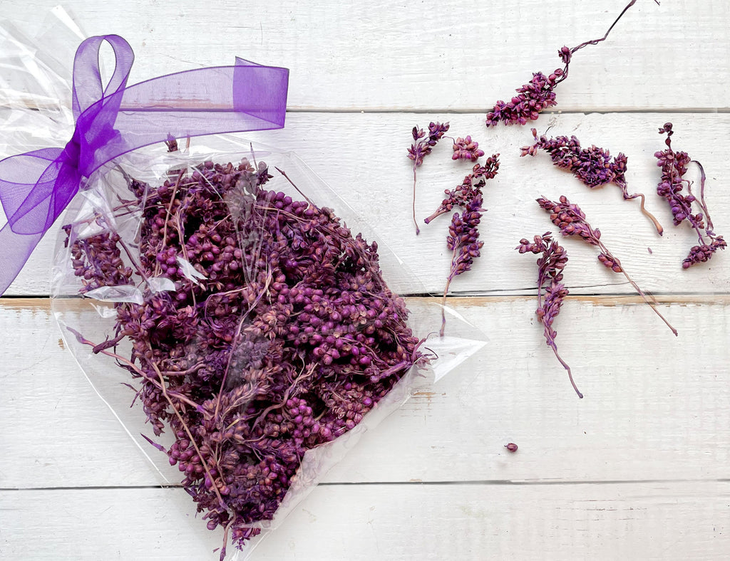 Dried purple flowers in a clear package with a purple ribbon on a white wooden surface.