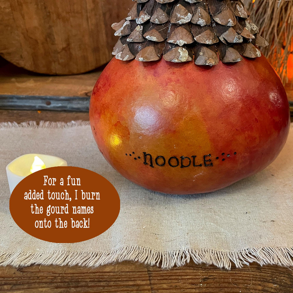Decorative gourd with a pinecone on top, placed on a textured surface with a candle and text overlay.