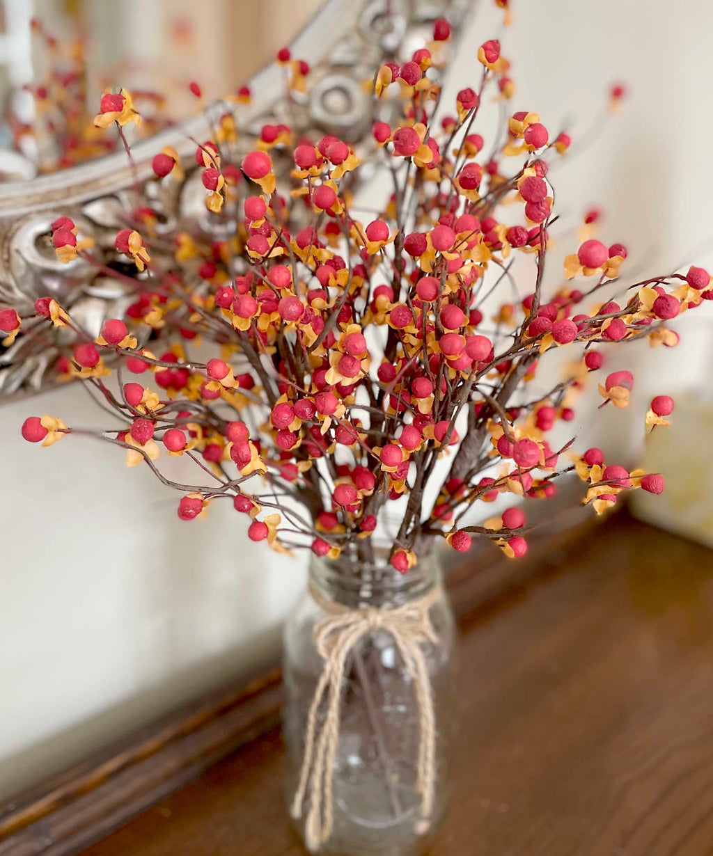 Decorative arrangement of red and orange berries in a glass vase with twine, against a neutral background.