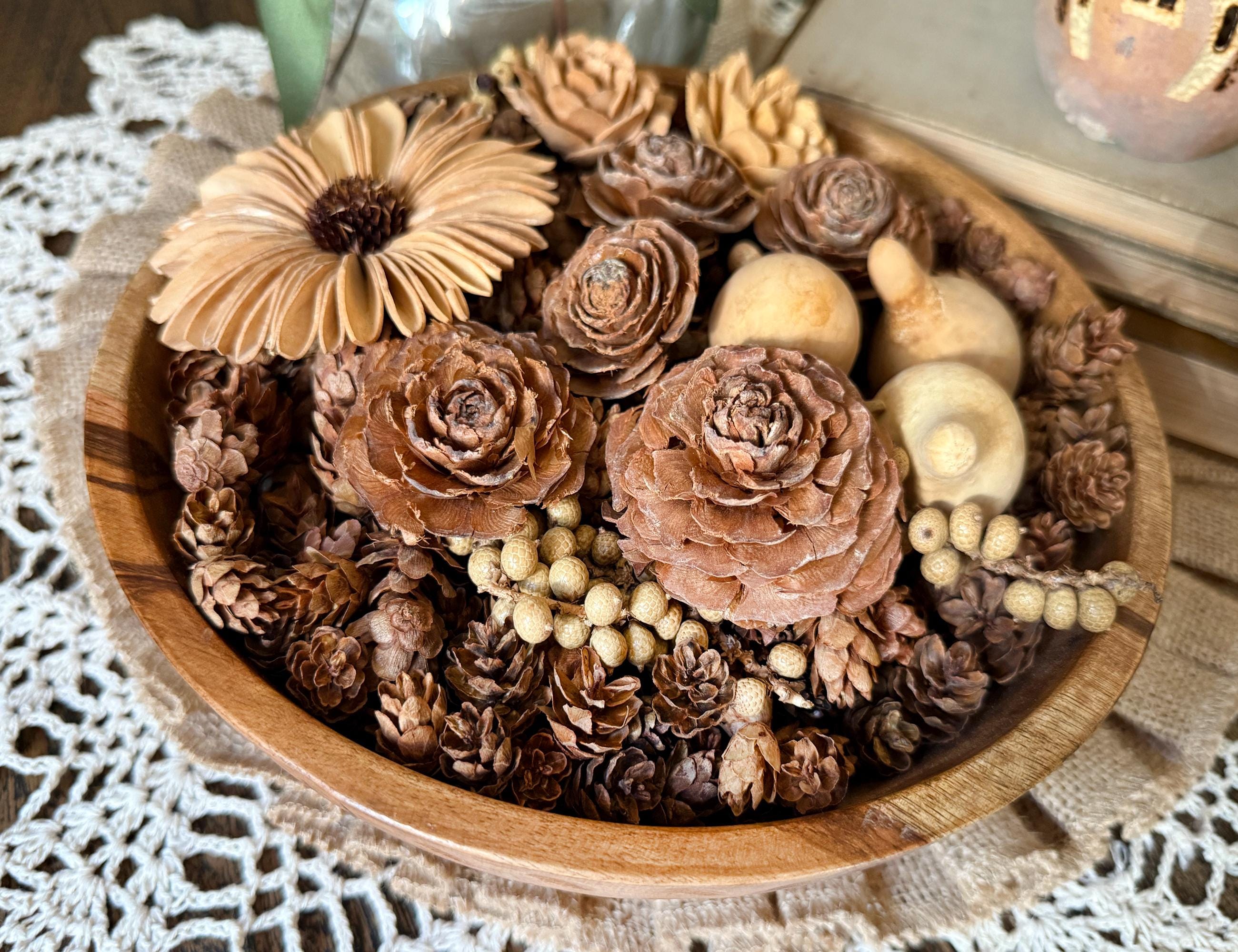 Wooden bowl filled with decorative pinecones and flowers on a lace tablecloth.