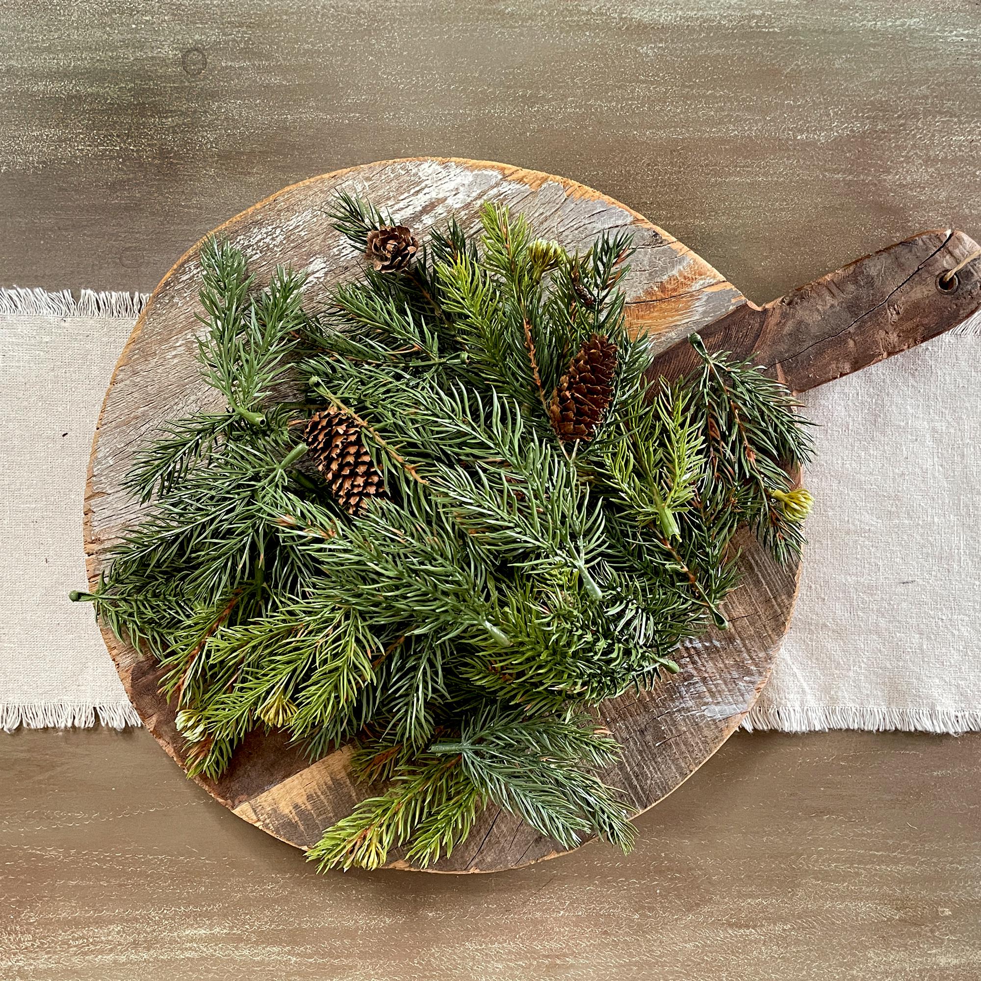 Wooden cutting board with a wreath of greenery and pine cones on a wooden surface.