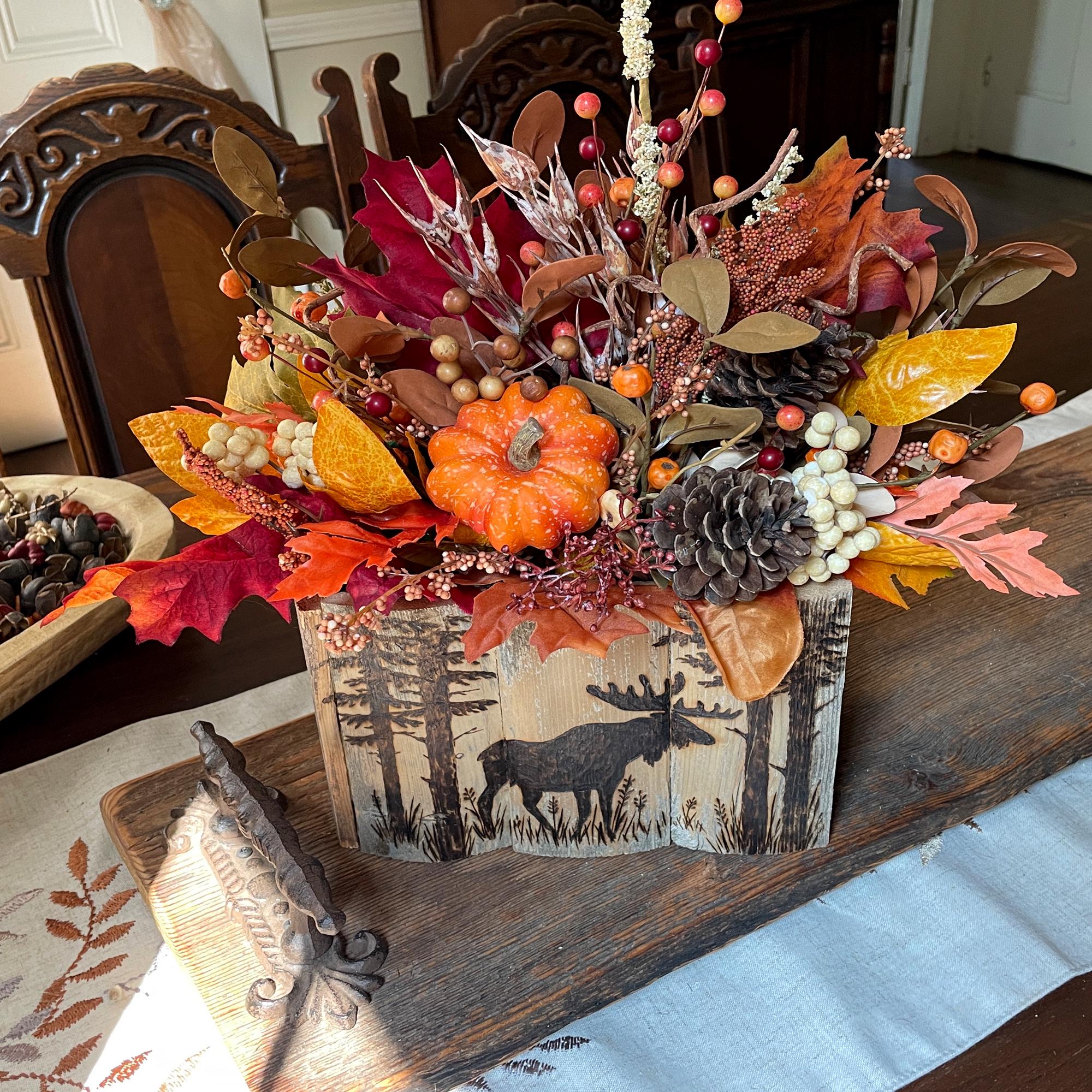 Autumn-themed floral arrangement in a wooden box with a deer design on a table.