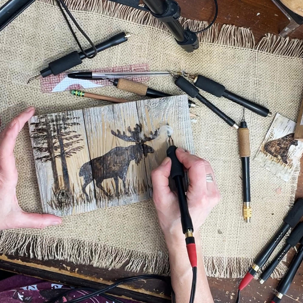 Person using a tool to create or modify a wooden plaque with a moose design on a textured surface.