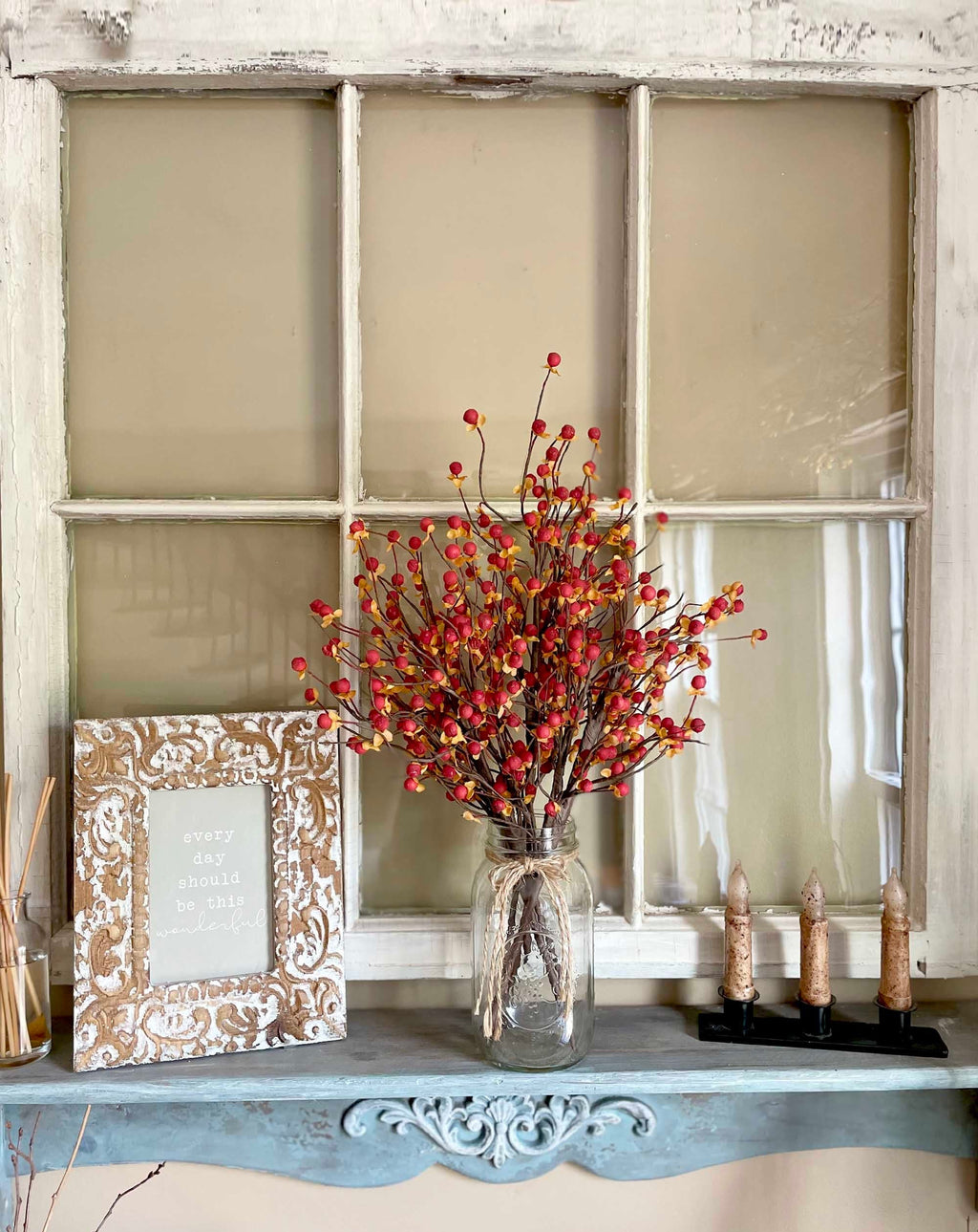 Decorative shelf with a vase of red berries, framed picture, and candles against a window pane background.