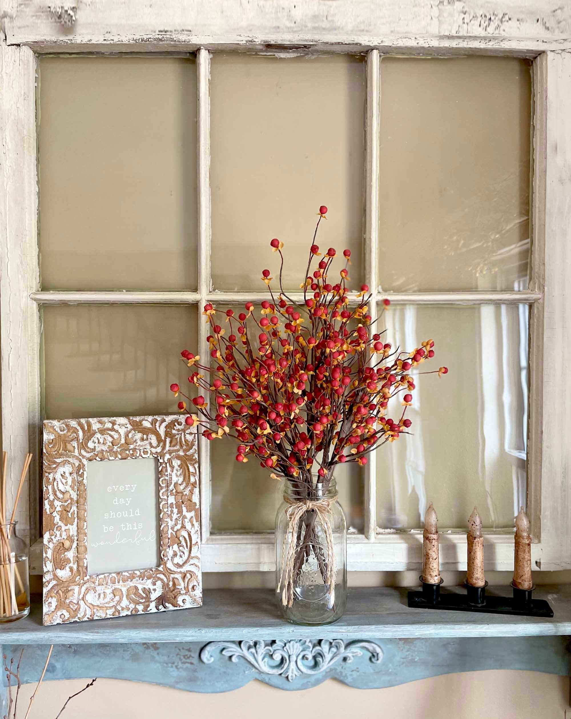 Decorative shelf with a vase of red berries, framed picture, and candles against a window pane background.