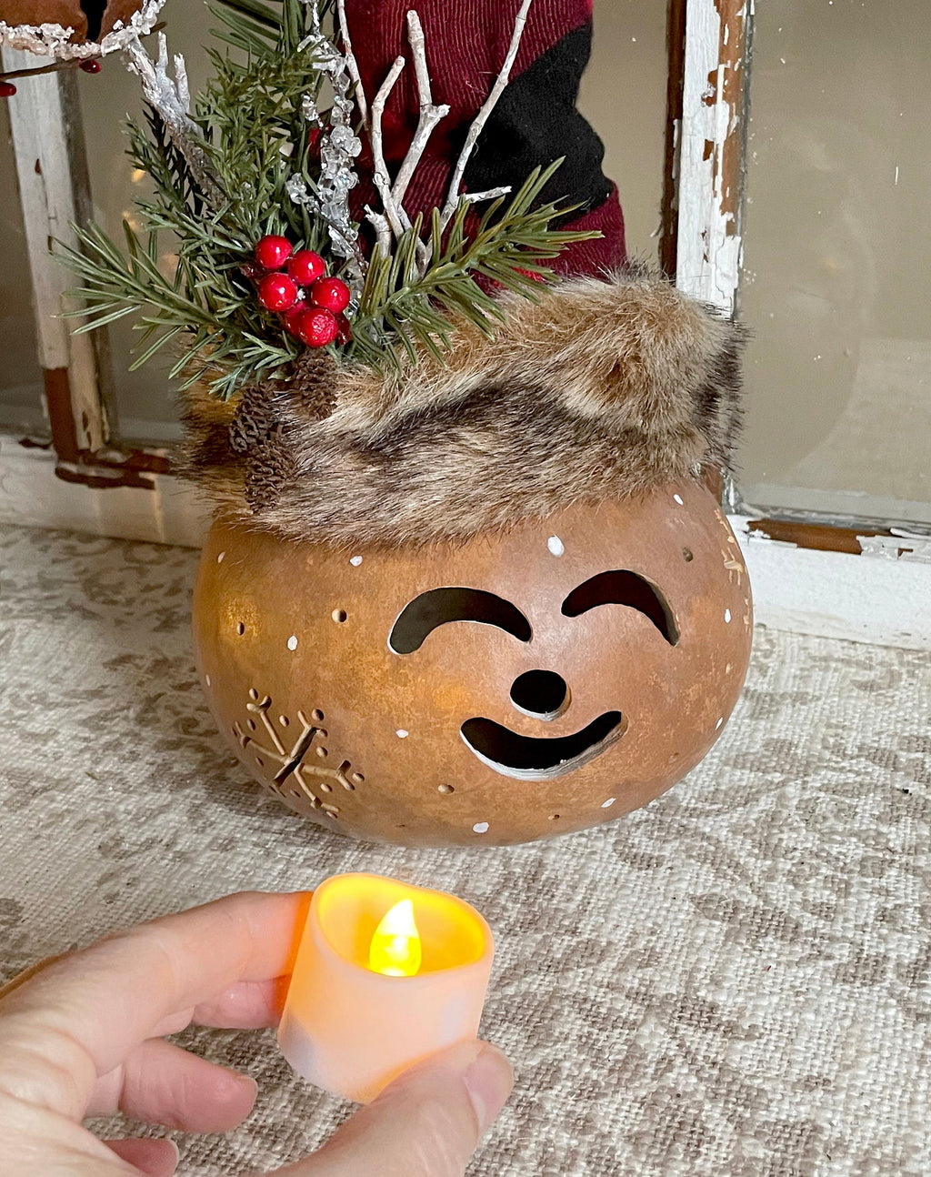 Decorative gourd with face cutout, fur top, and greenery on a textured surface.