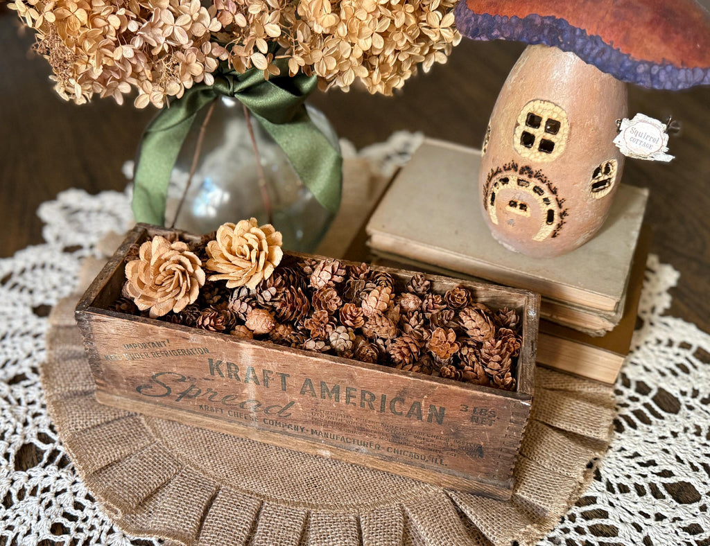 Decorative setting with a wooden box filled with pinecones, flowers, and a small house ornament on a lace tablecloth.